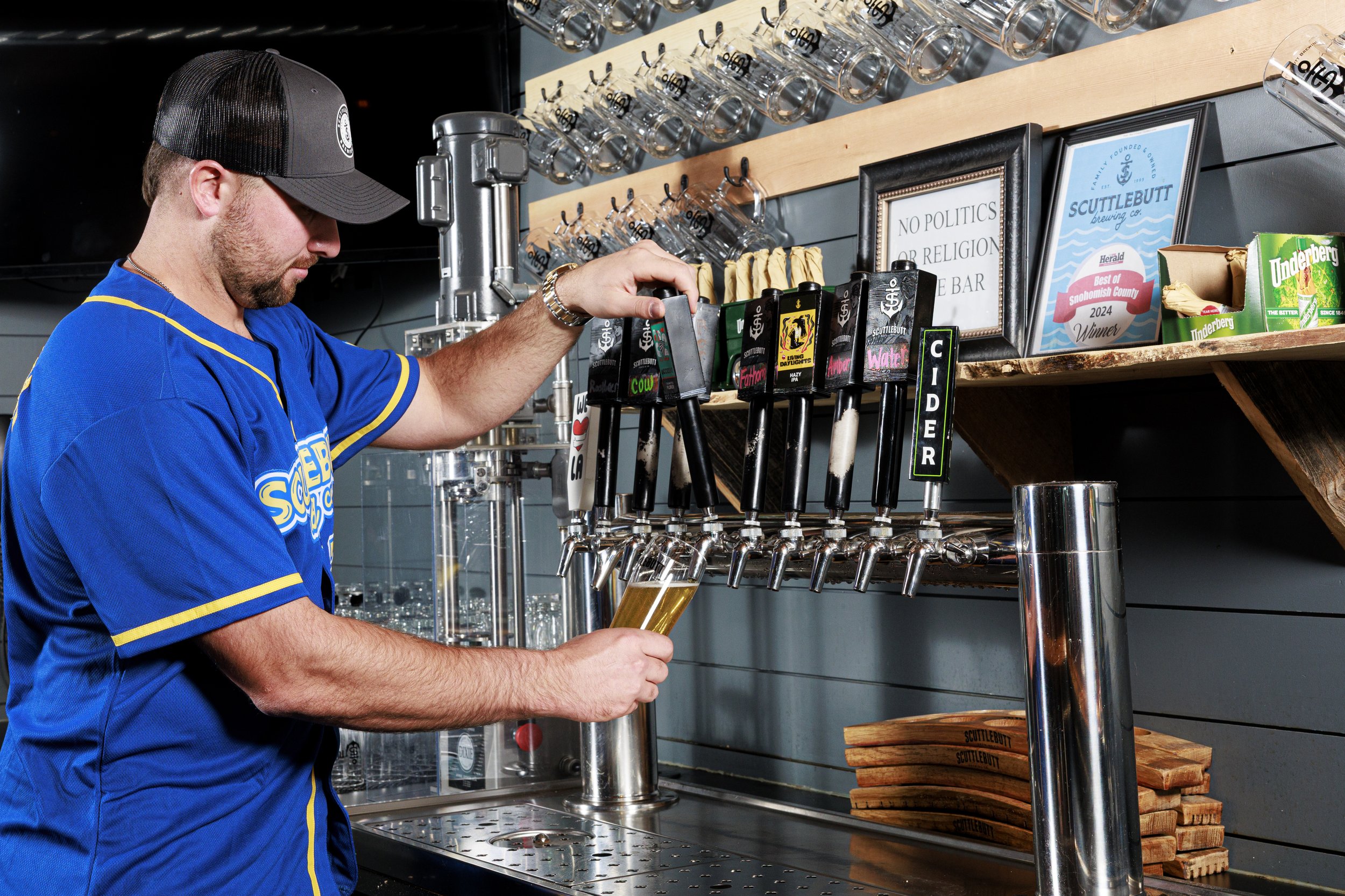 Cal Raleigh in a blue shirt and black baseball cap pouring beer from a tap at a bar with shelves of glassware, framed signs, and snack items in the background.