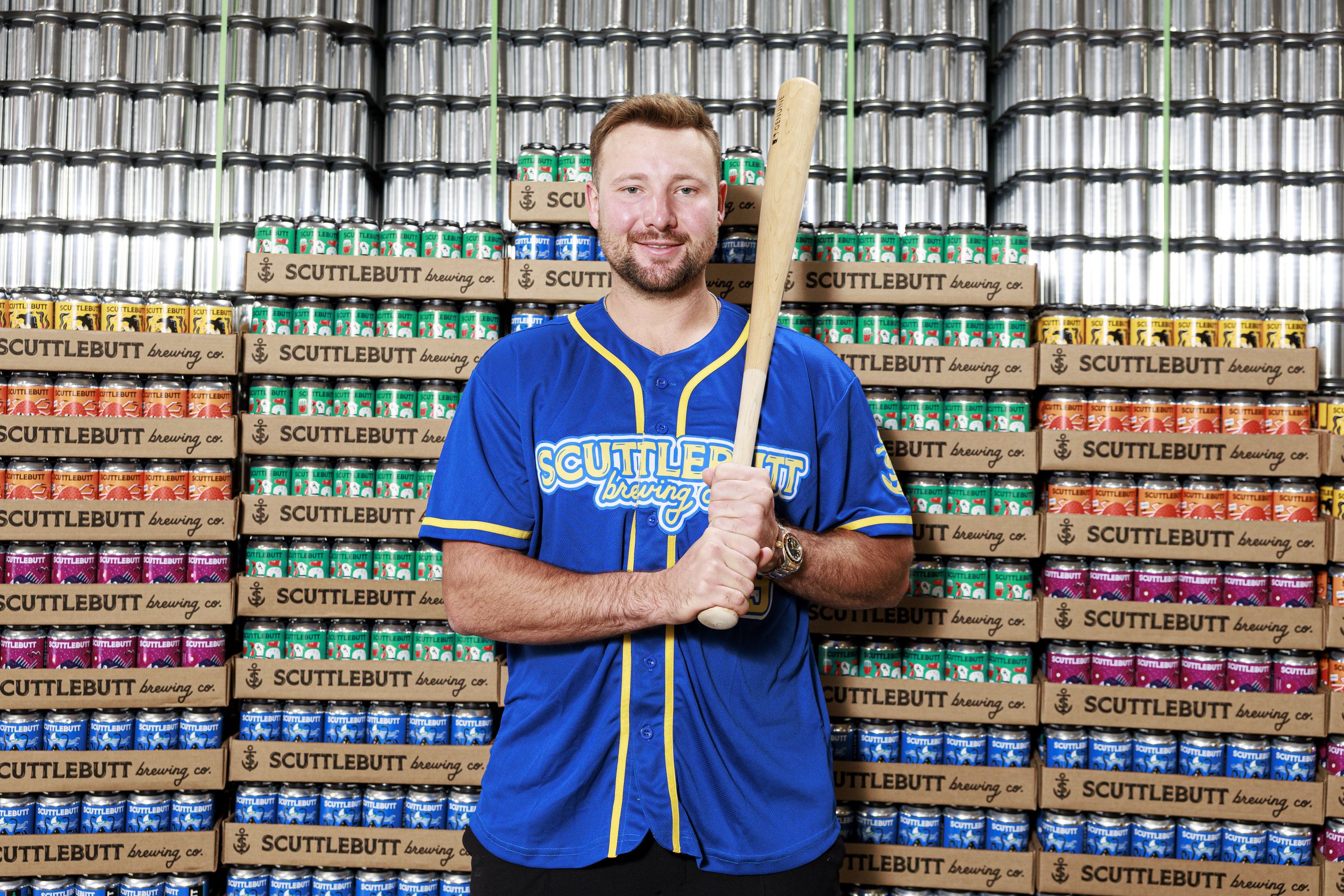 A man in a blue and yellow sports jersey standing in front of a wall of colorful beer cans with categories such as 'Littlebutt Brewing Co.' and holding a wooden baseball bat.