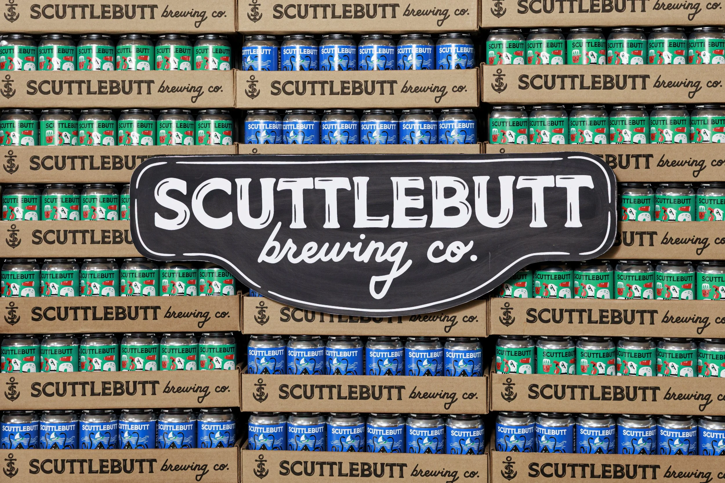 Rows of cans with unicorn and aquatic designs, stacked on shelves with cardboard boxes labeled 'Scuttlebutt Brewing Co.' and a large chalkboard-style sign reading 'Scuttlebutt Brewing Co.' in front.
