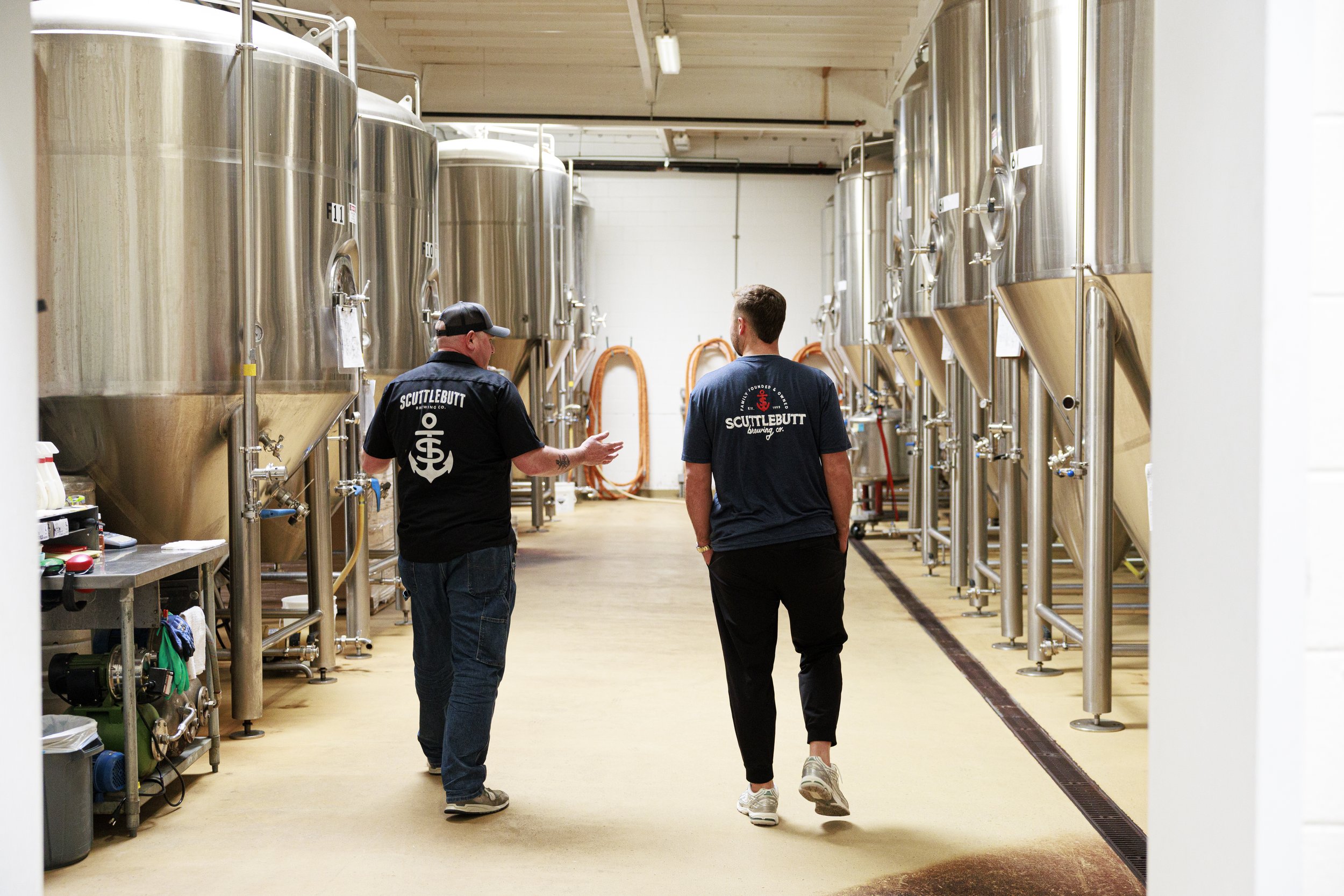 Two men walking and talking inside a brewery, surrounded by large stainless steel fermentation tanks and brewing equipment.