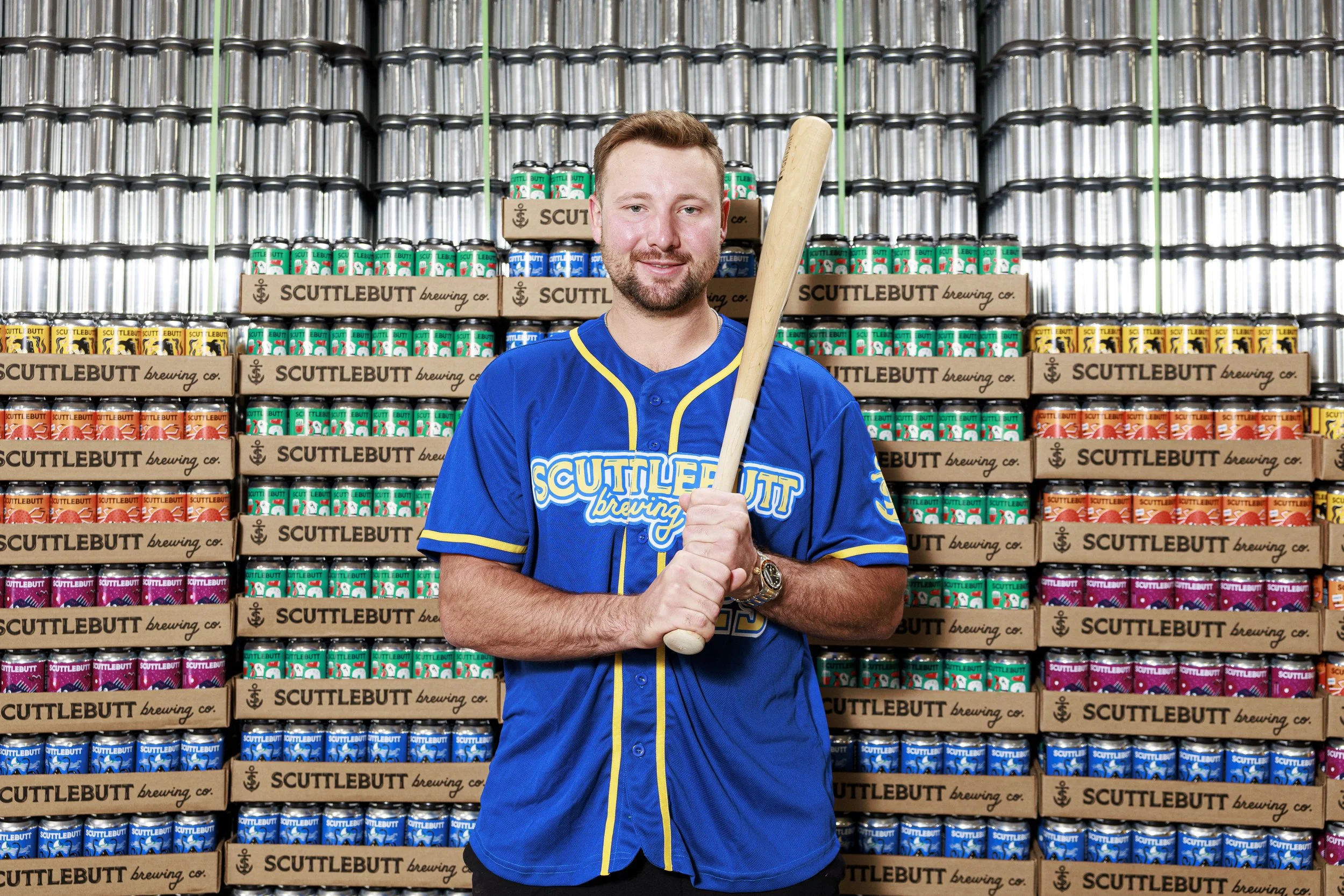 A man in a blue sports jersey stands in front of shelves stacked with cans of beer. He holds a baseball bat and smiles at the camera.