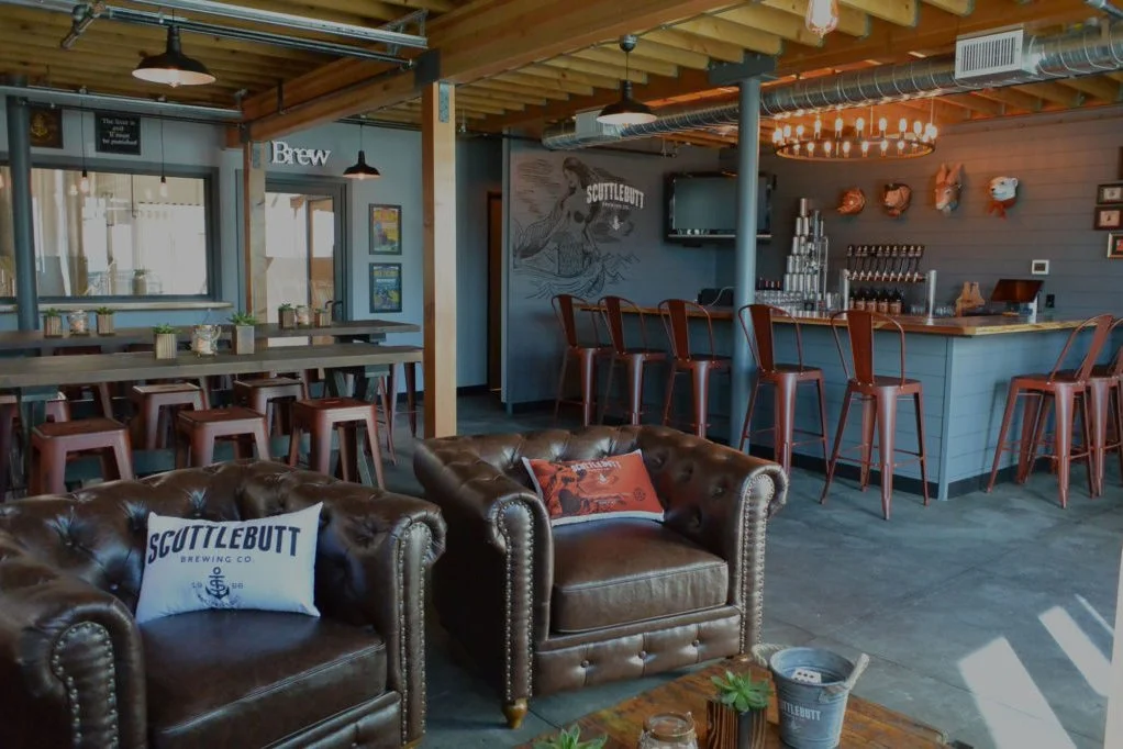 Interior of a cozy brewery with leather armchairs, a bar area with high chairs, a high table with chairs, and wall decorations.