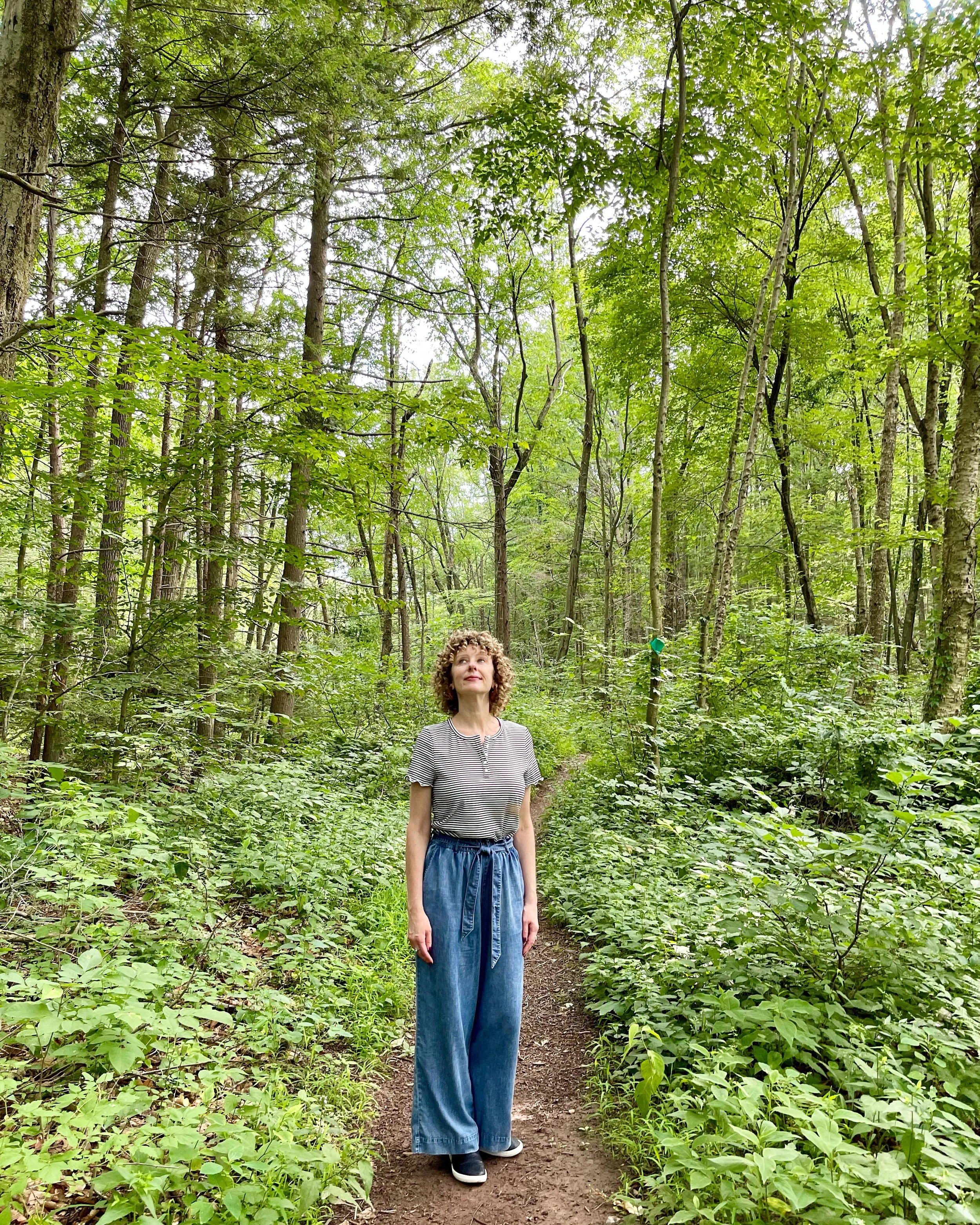 A woman with curly hair in a striped shirt and high-waisted wide-leg jeans walking on a forest trail surrounded by green trees and bushes.