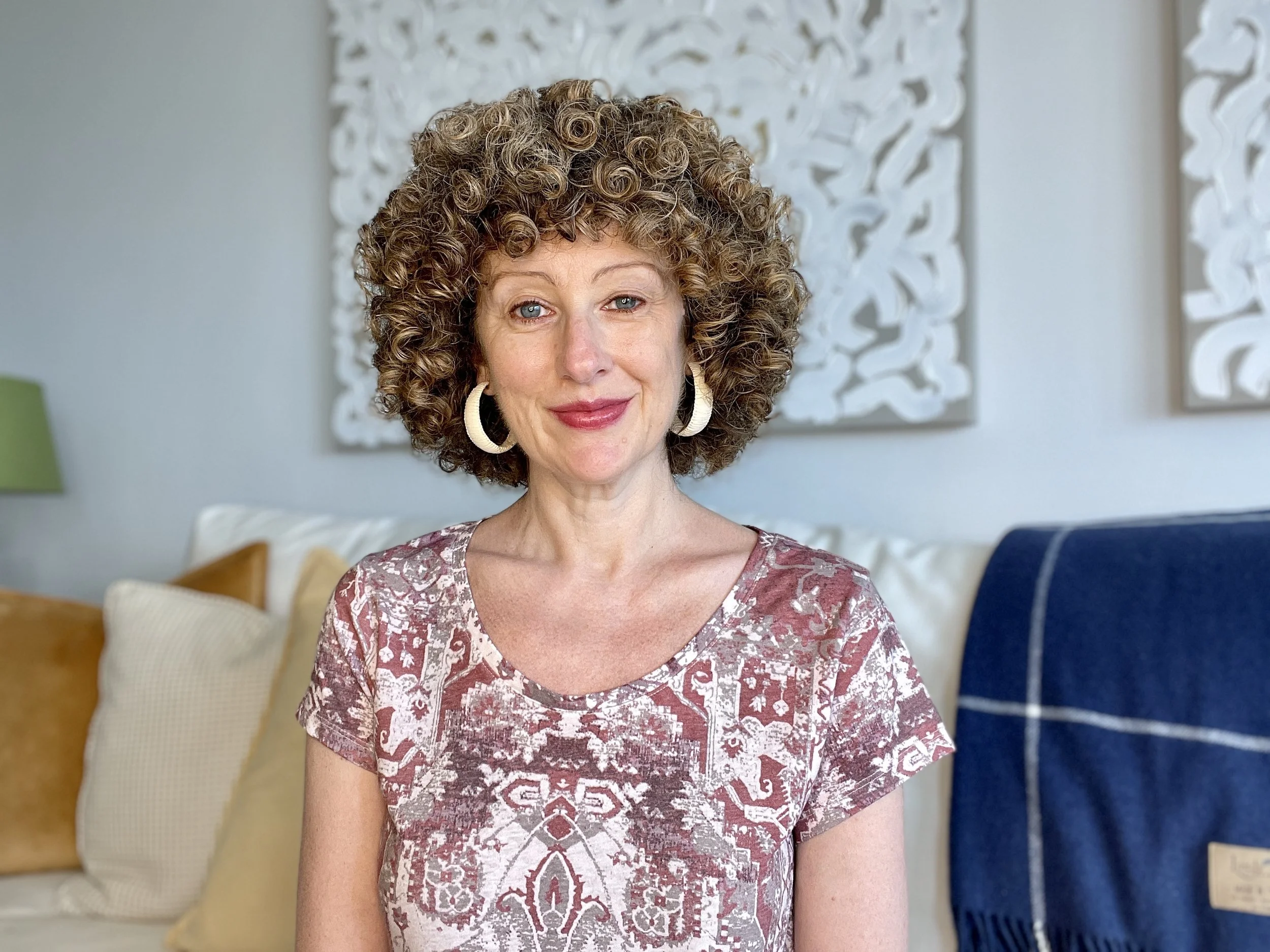 A woman with curly hair and hoop earrings smiling in a living room with colorful pillows and decorative wall art.