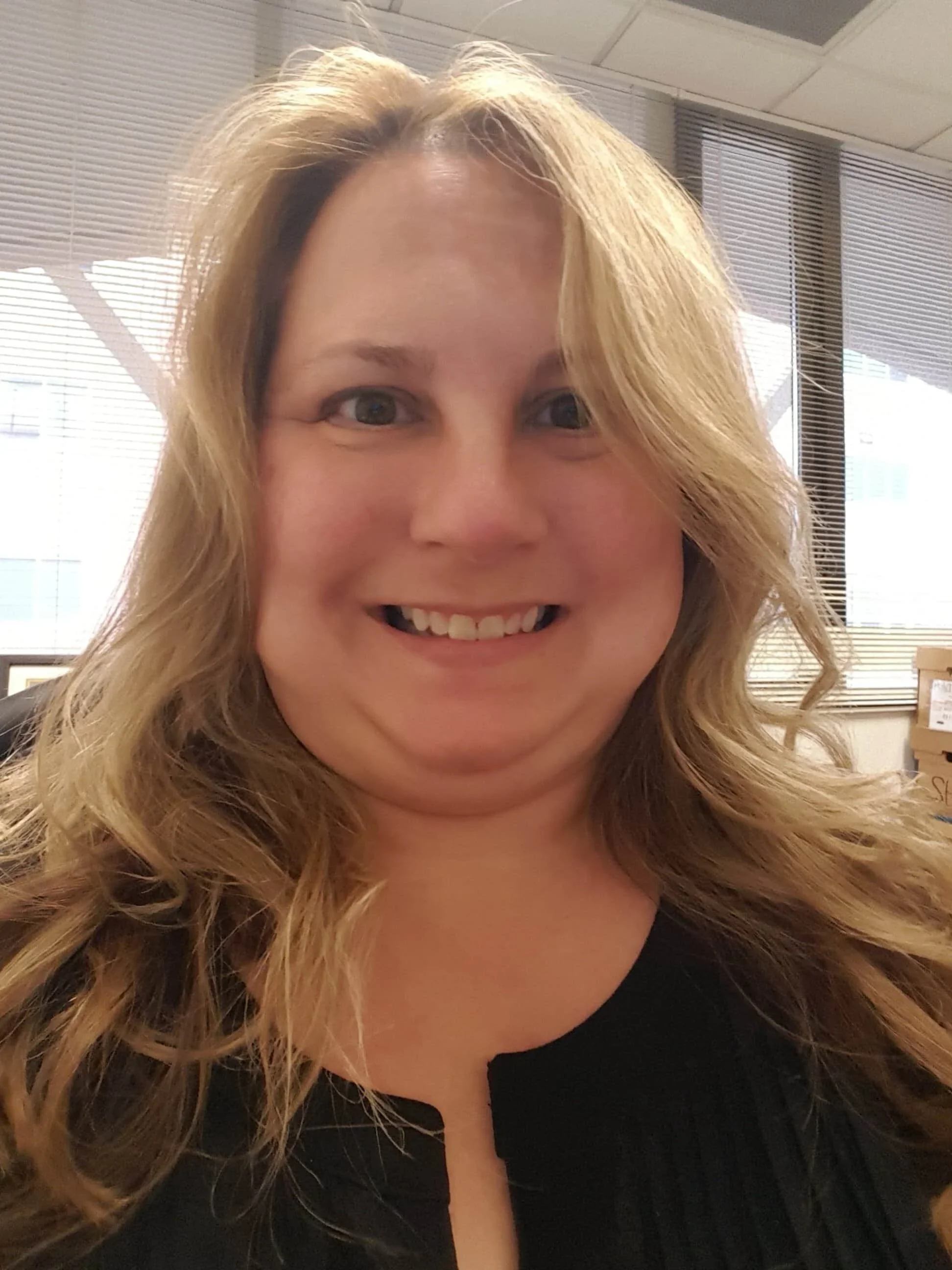 A woman with wavy blonde hair smiling at the camera in an indoor office environment with windows, blinds, and office supplies in the background.