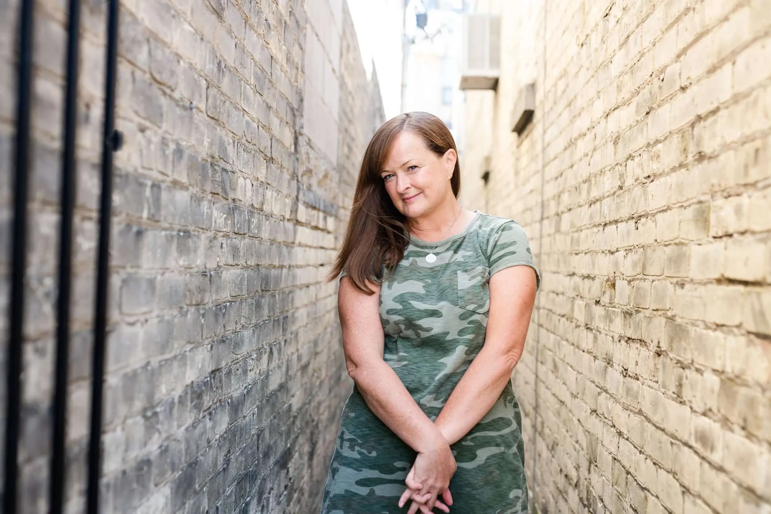 A woman with long brown hair wearing a green camouflage t-shirt, standing in a narrow alley between two brick walls.