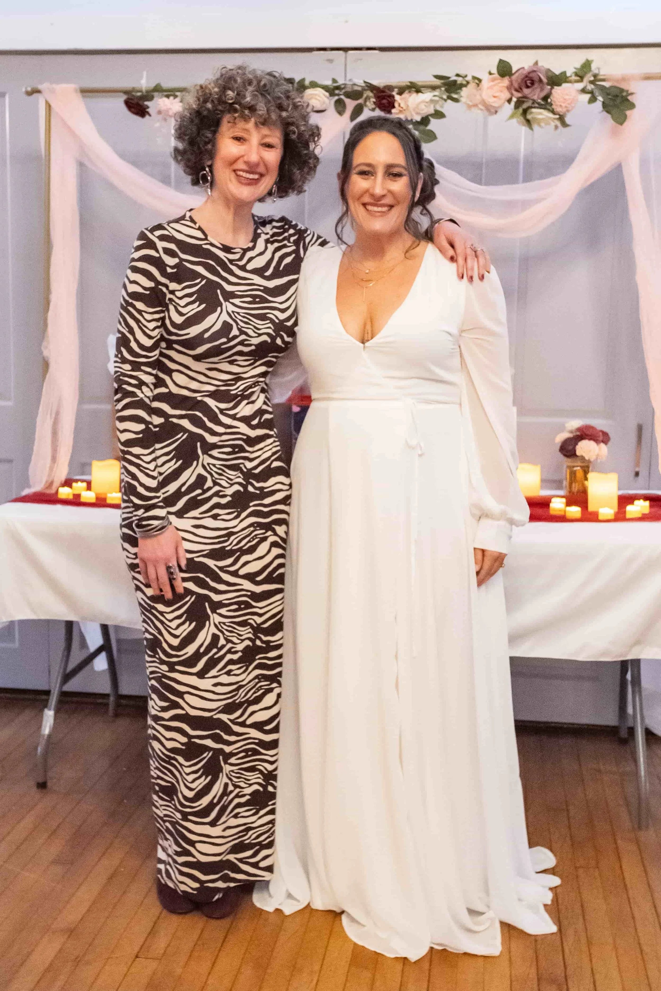 Two women smiling and posing together at a celebration. One is dressed in a zebra-print long dress, and the other is wearing a white dress with long sleeves, a deep V-neck, and a flowing hem. They are standing in front of a decorated table with candles and flower arrangements.
