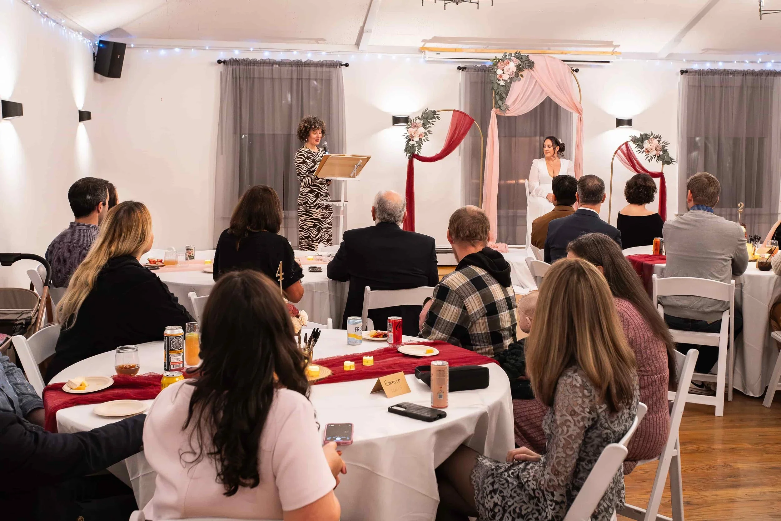 People seated at round tables watching a woman speak on a decorated stage with floral arrangements and pink drapery in a banquet hall.