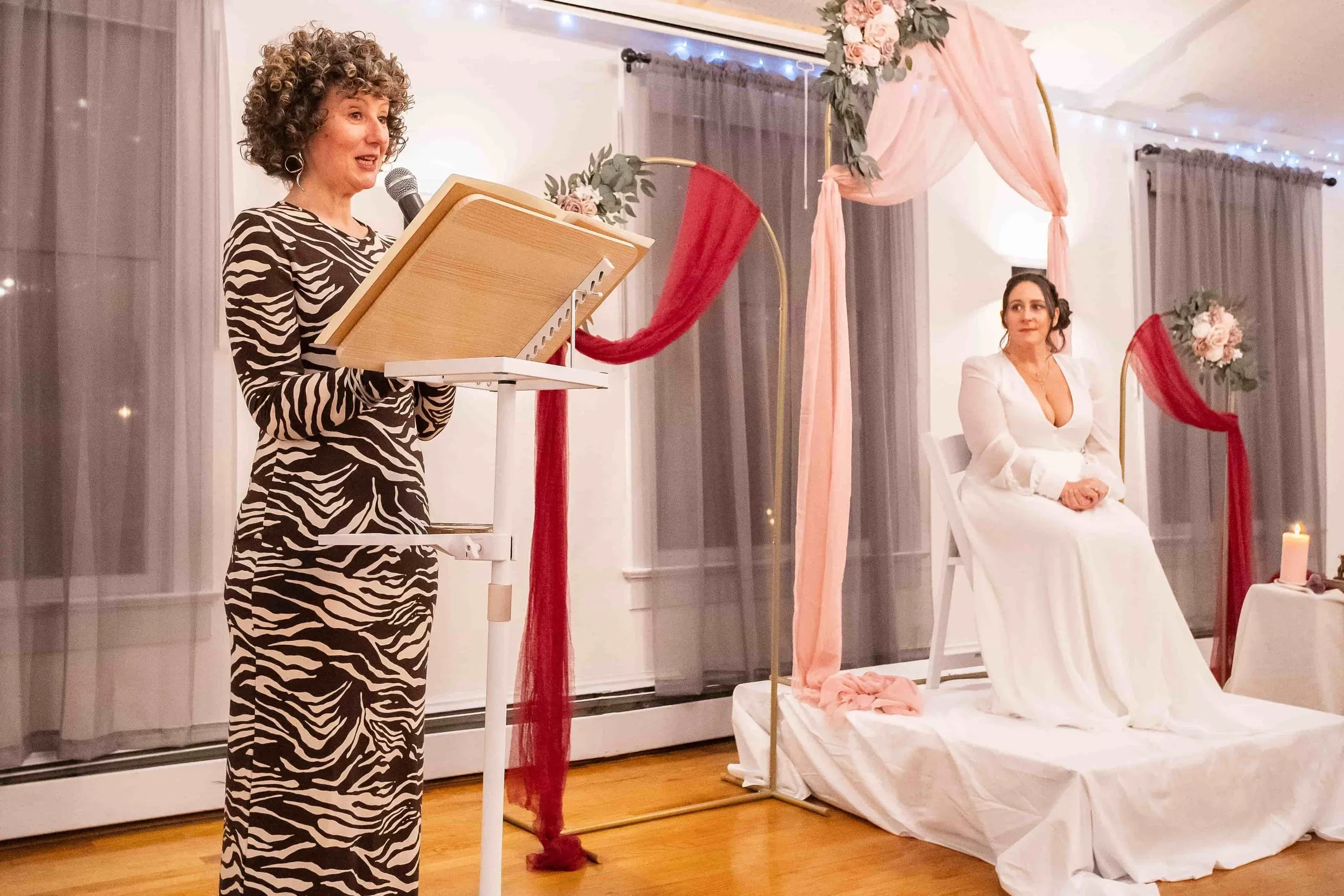 A woman giving a speech at a wedding reception. The bride, dressed in a white gown, is seated on a chair decorated with pink and red fabric and flowers. There are fairy lights and curtains in the background.