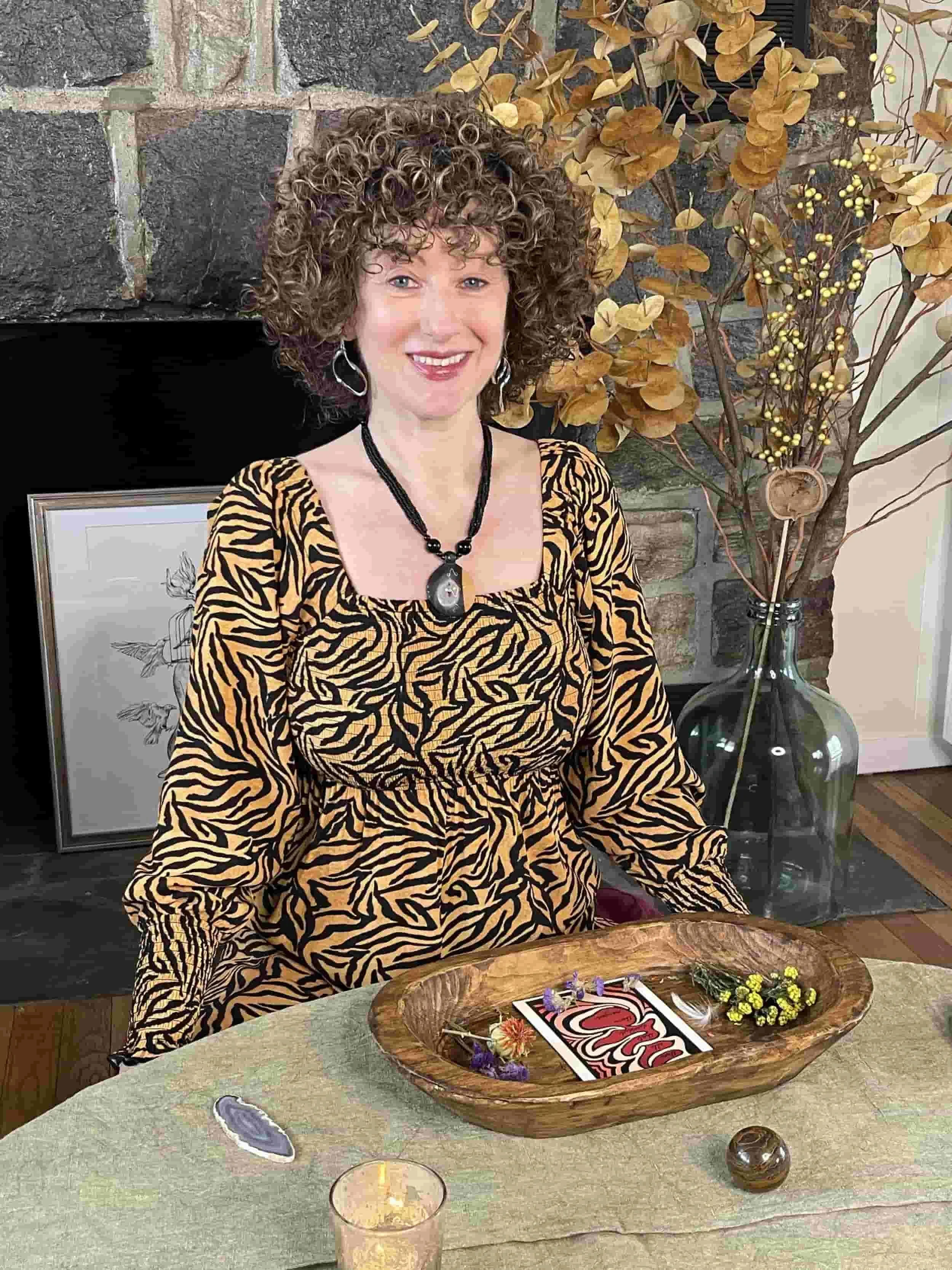 A woman with curly hair wearing a tiger print dress, jewelry, and makeup, sitting at a table with a decorative wooden bowl, flowers, and a candle, in a room with a brick fireplace and rustic decor.