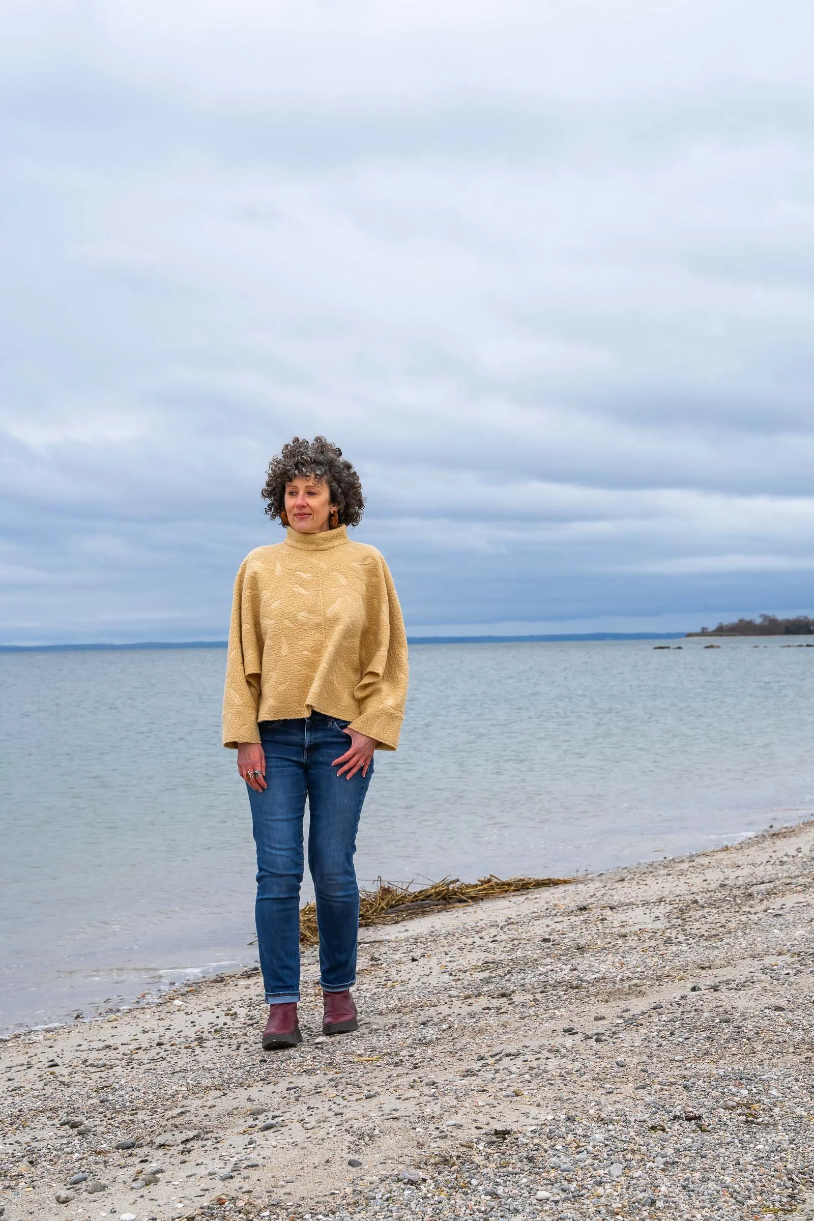 A woman with curly hair wearing a beige sweater, blue jeans, and boots standing on a beach near a body of water under a cloudy sky.
