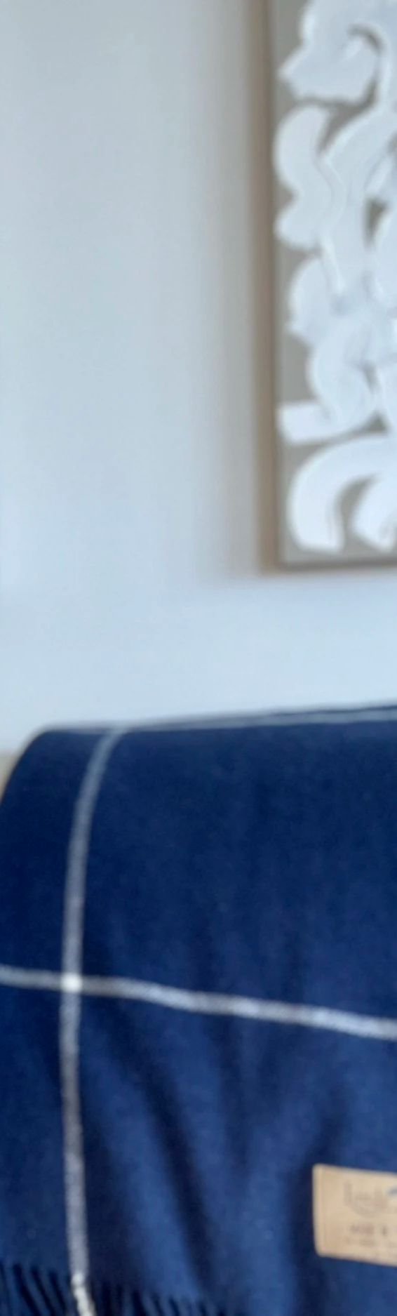 Close-up of a blue fabric surface with white stitching in the foreground, partially showing a brown label, against a background of a light-colored wall and a framed wall art piece with white abstract design.