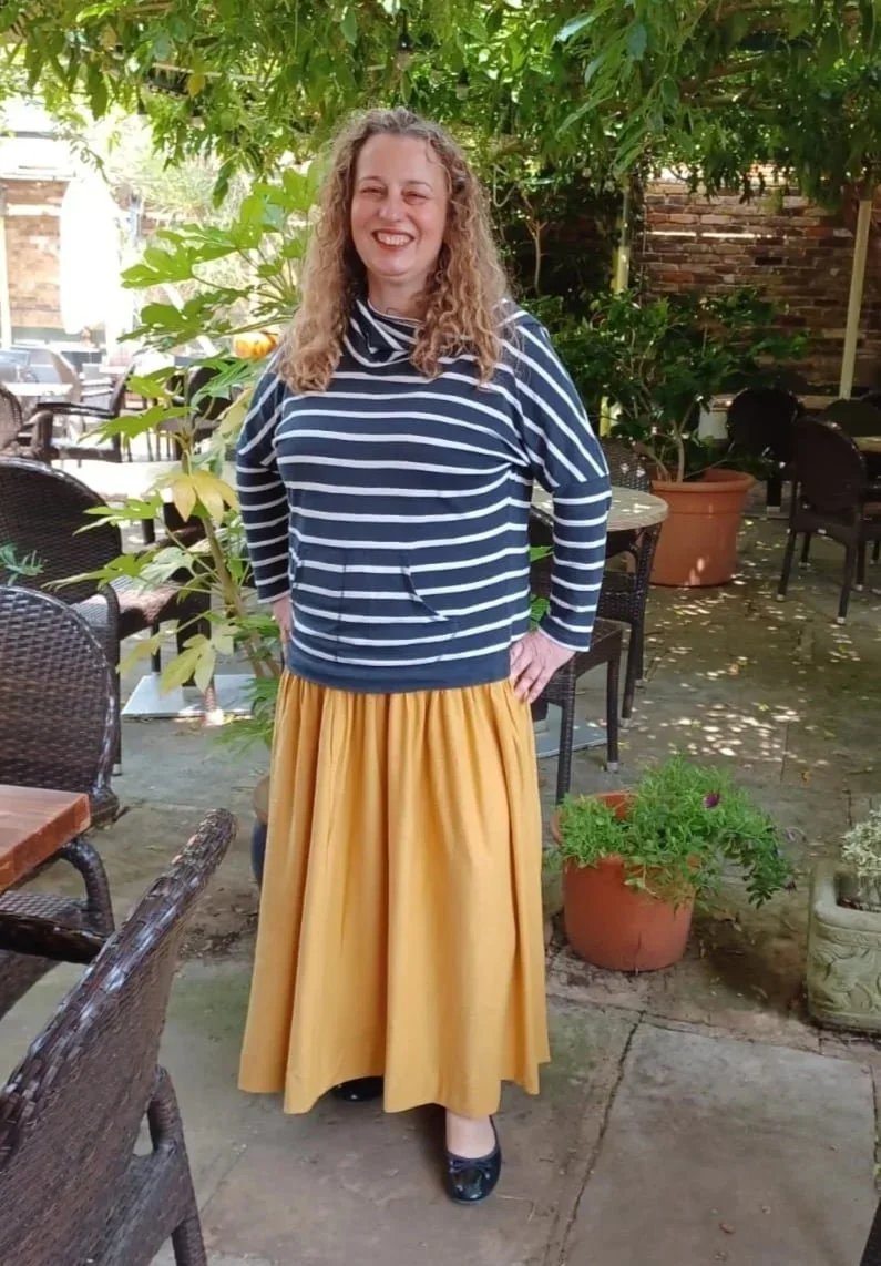 Smiling woman with curly hair in a striped navy and white shirt, yellow skirt and black shoes standing outdoors in a garden setting with potted plants and patio furniture.