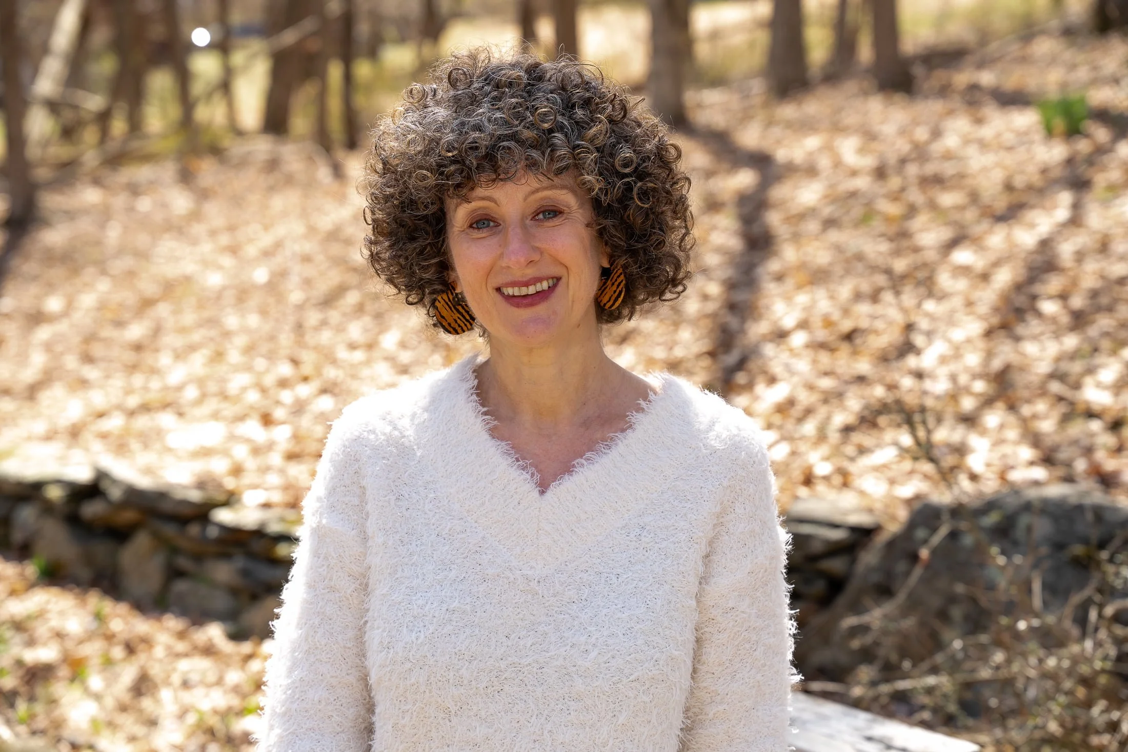 A woman with curly hair wearing a white textured sweater and striped earrings standing outdoors in a sunlit forested area.