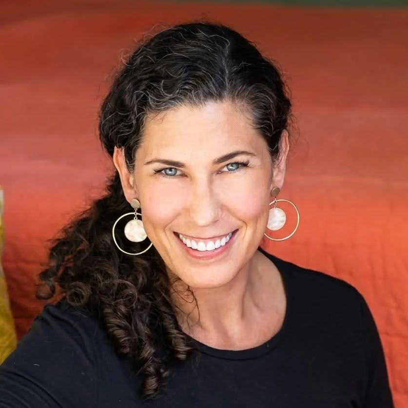 Close-up of a woman with curly dark hair, blue eyes, and a bright smile wearing large hoop earrings and a black top, sitting outdoors against an orange background.