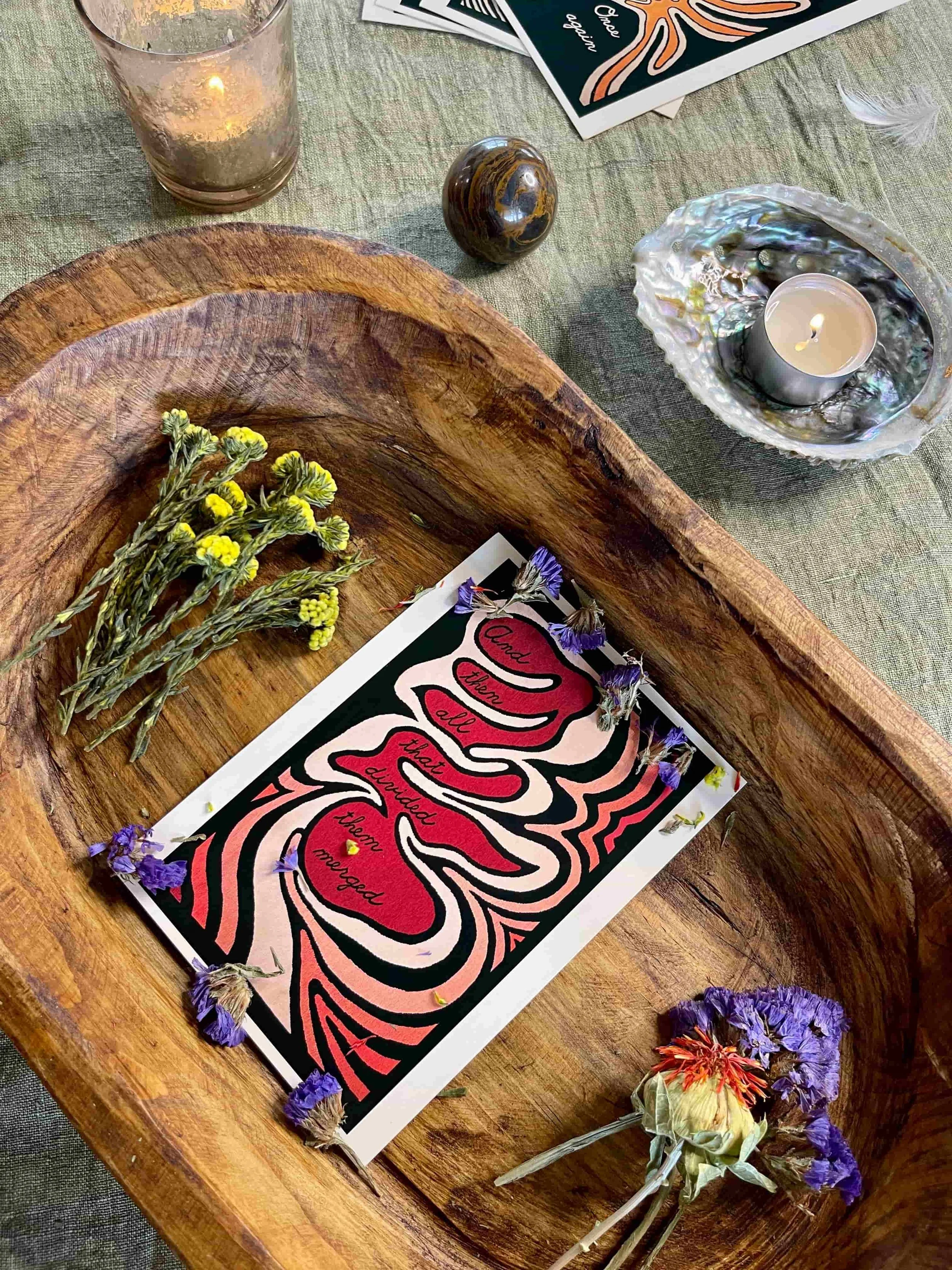 A wooden tray with dried flowers, a colorful card with abstract patterns and handwritten message, surrounded by glass candles, a polished stone, and scattered flowers on a green tablecloth.