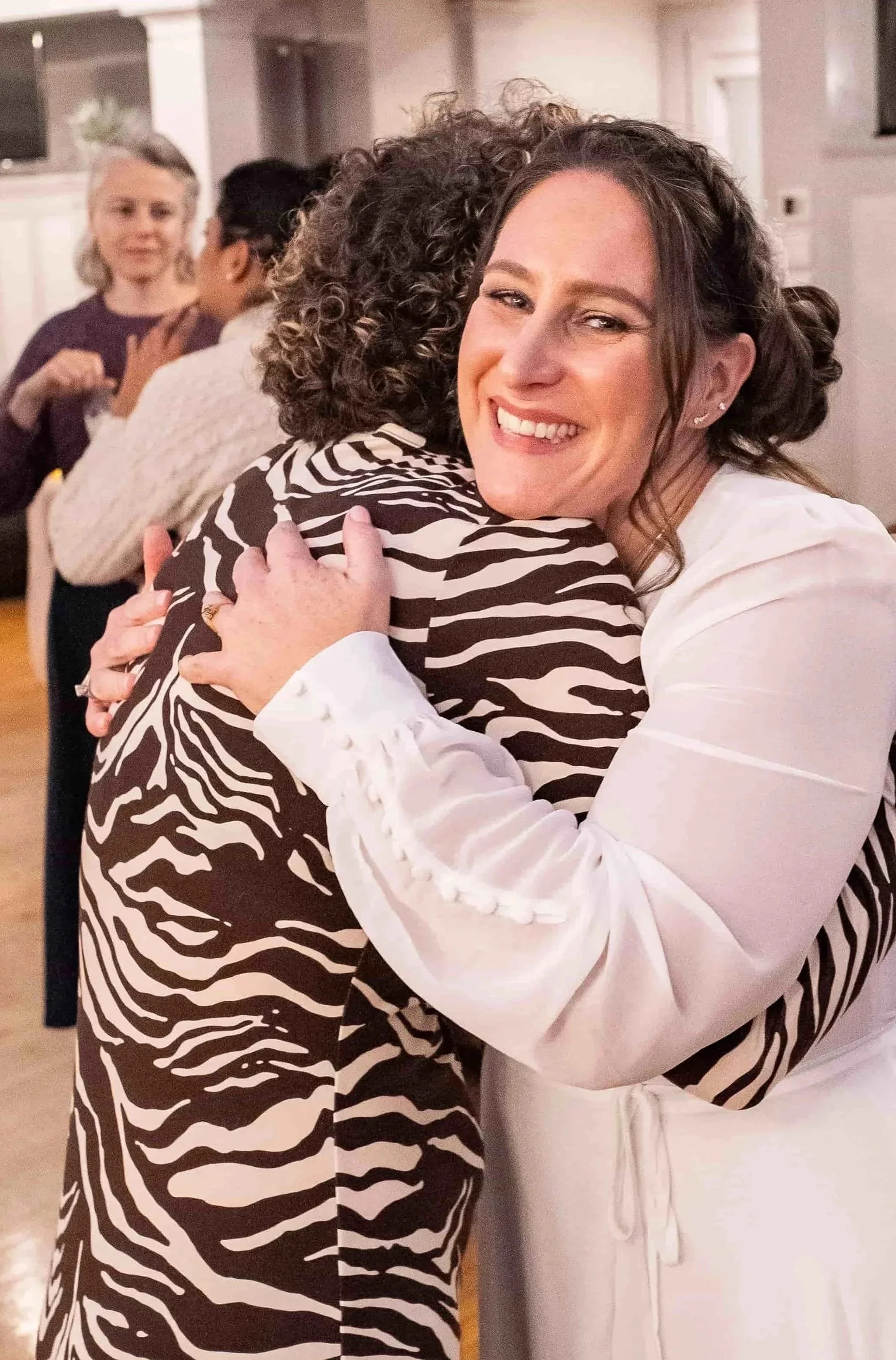 Two women hug warmly at a social gathering, smiling. Other people are in the background, talking and mingling.