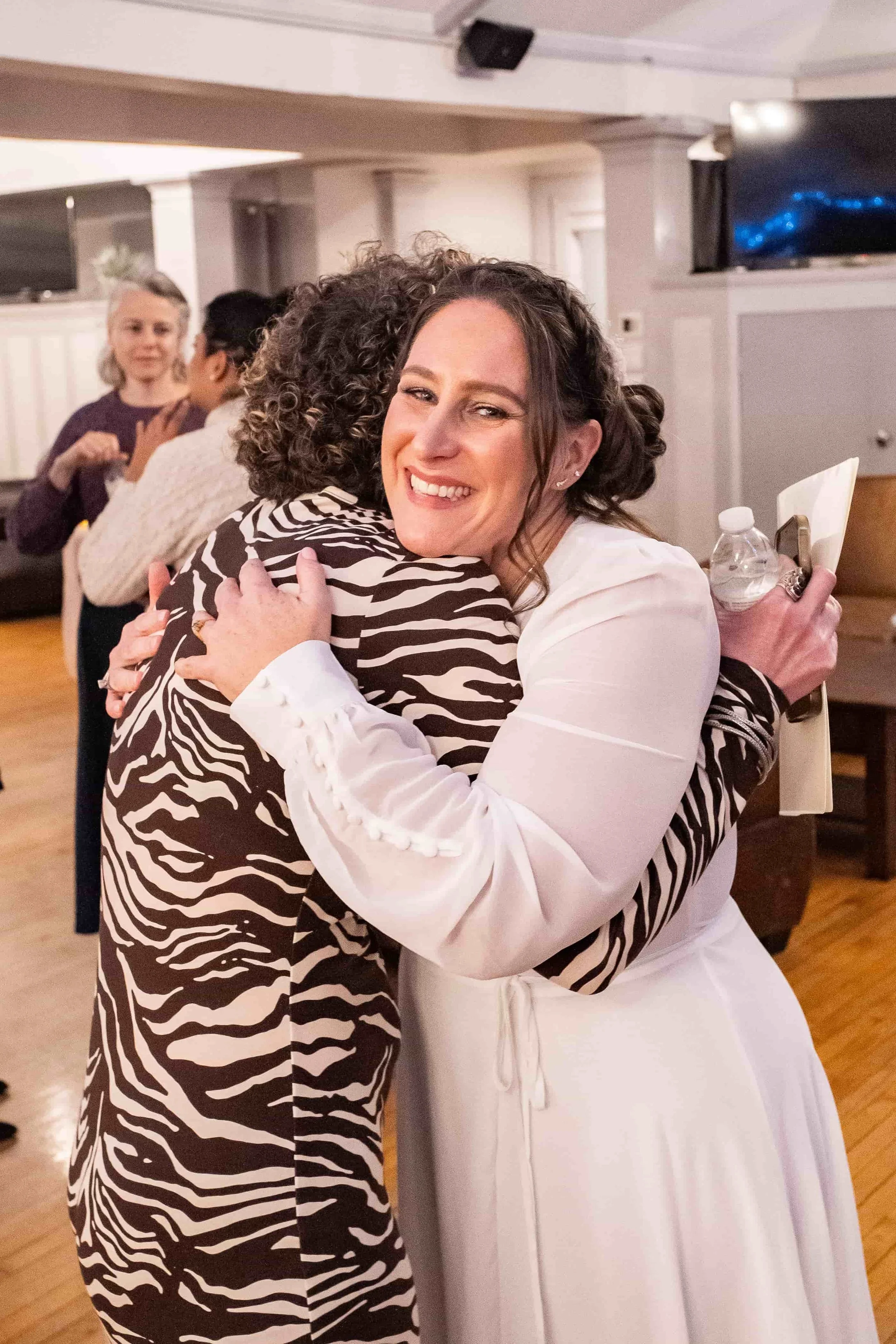 Two women hugging warmly at an indoor gathering, with others in the background.