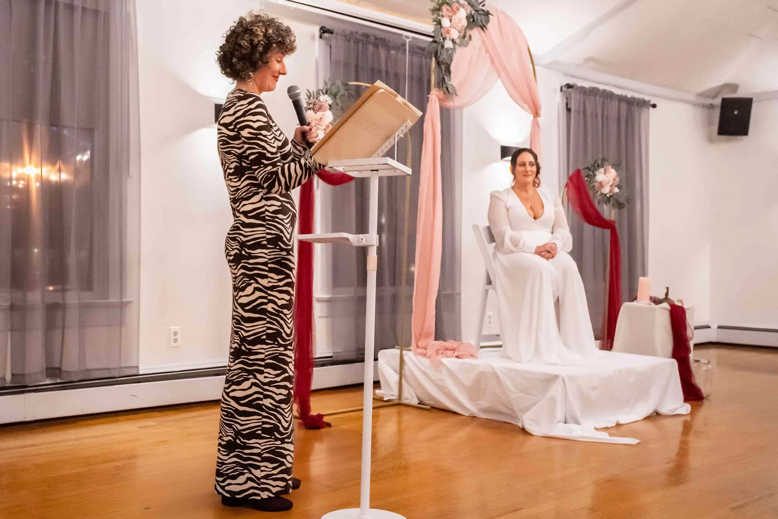 A woman with curly hair in a zebra print dress giving a speech at a wedding reception, standing behind a podium with a microphone. A bride in a white dress seated on a decorated platform with pink and red fabric and floral arrangements.