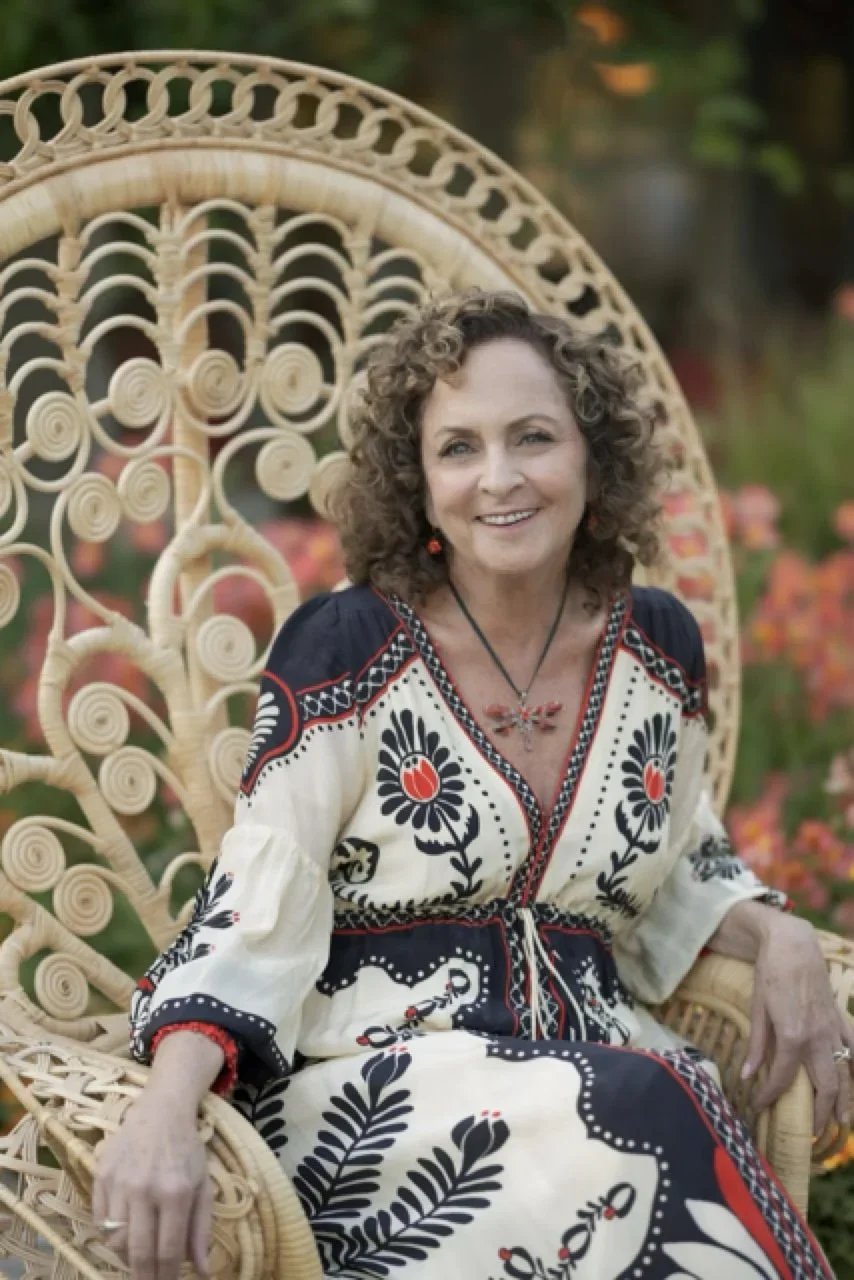 A woman with curly hair sitting on a decorative wicker chair in a garden, wearing a white dress with black and red floral patterns.