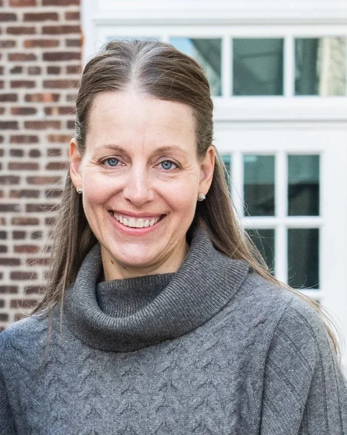 A woman with long light brown hair smiling, wearing a gray turtleneck sweater, standing outside in front of a brick wall and a white-framed window.