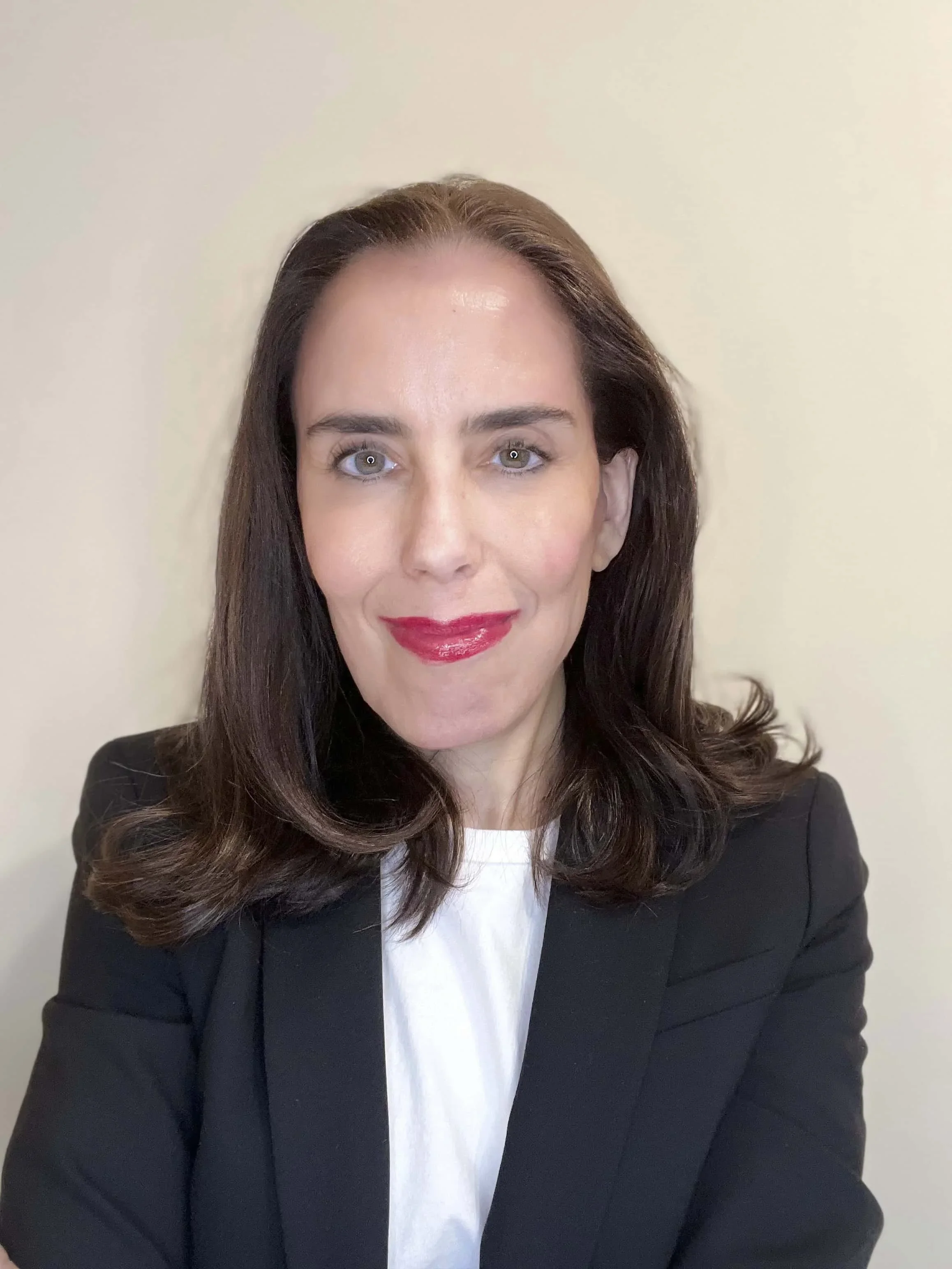 A woman with brown hair wearing a black blazer and white shirt, smiling with red lipstick, against a plain light-colored wall.