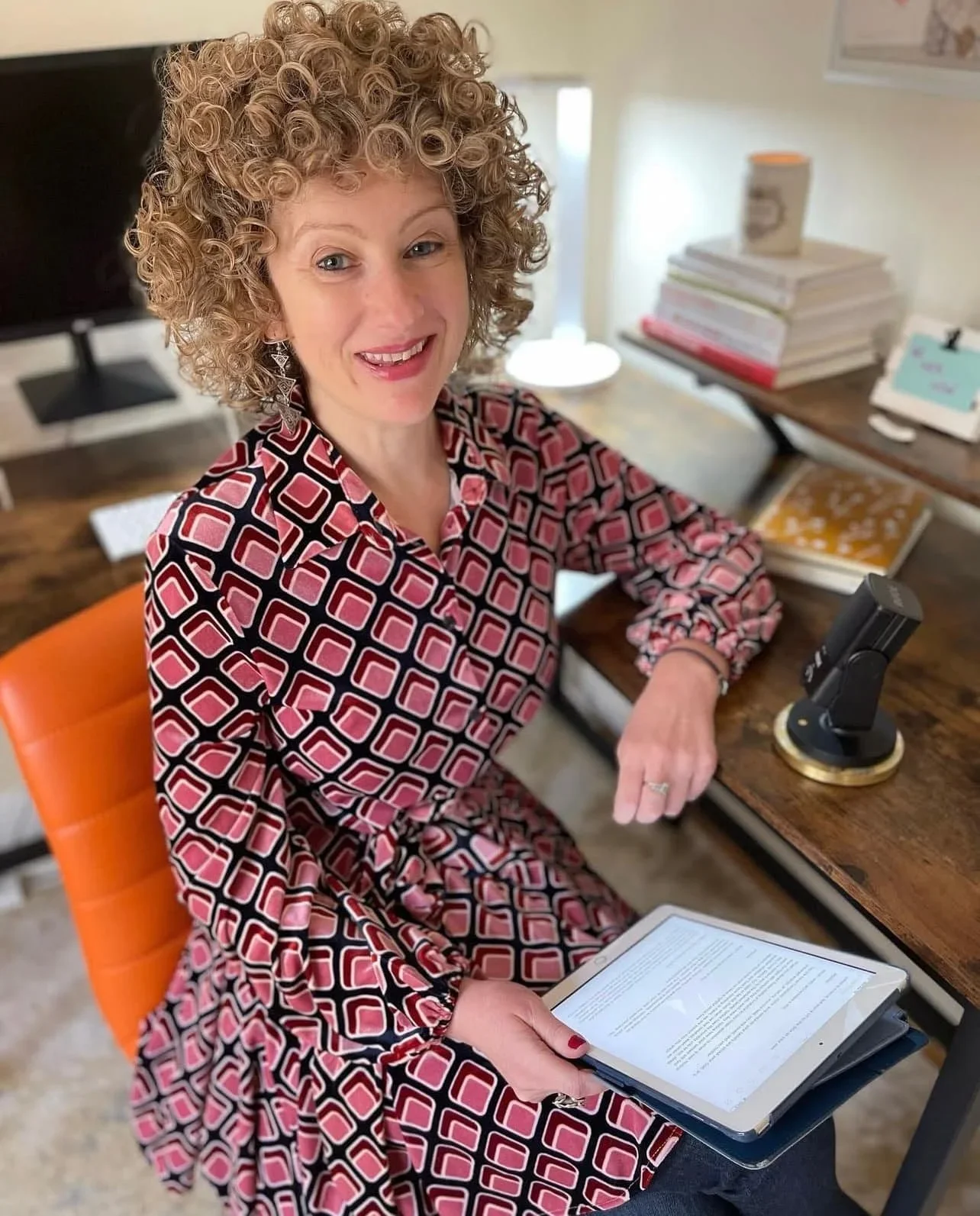 A woman with curly blonde hair sitting at a desk, holding an e-reader, smiling at the camera. The desk has stacks of papers, a computer monitor, and a microscope.