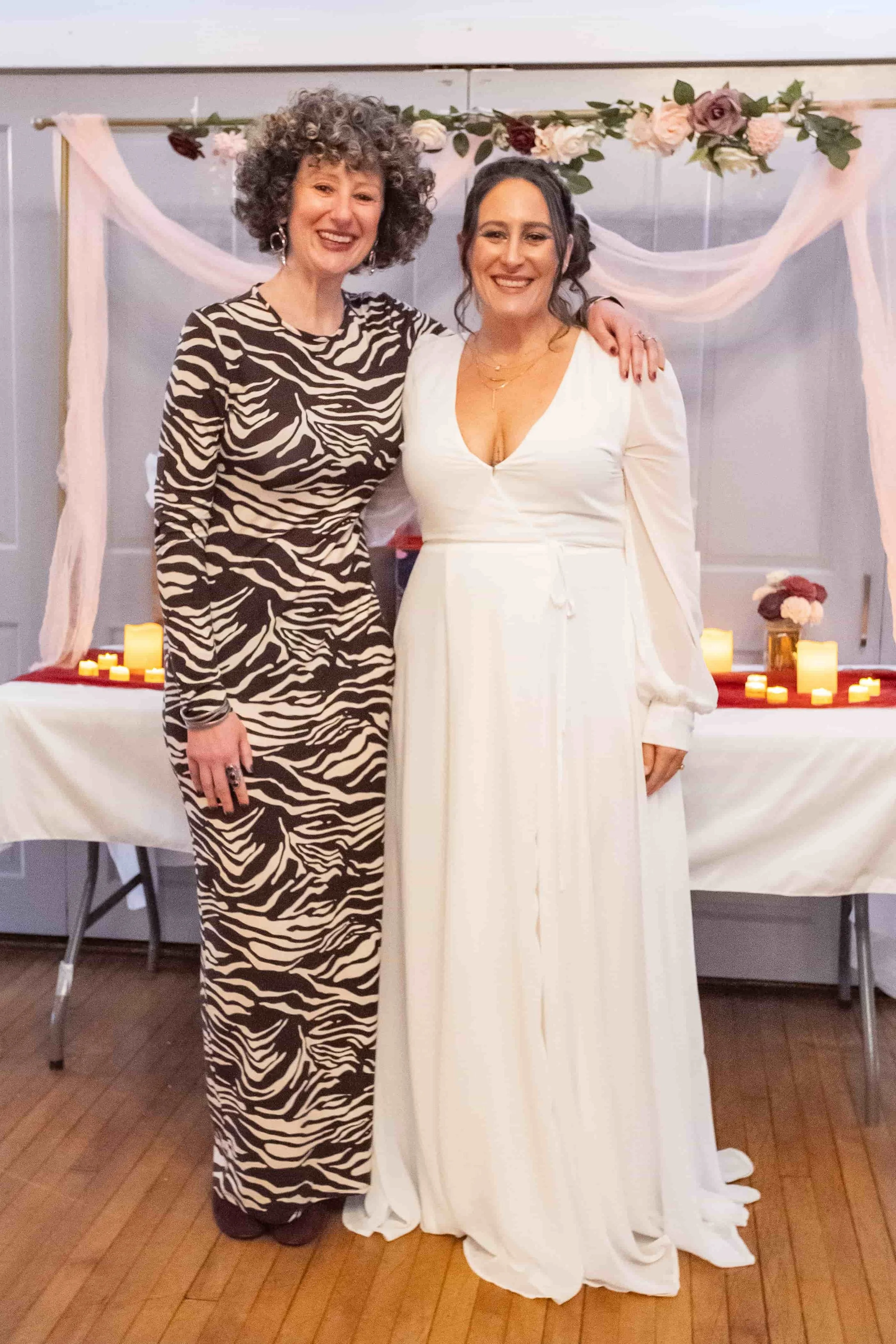 Two women standing together at what appears to be a celebration or reception. One woman in a zebra print dress and the other in a white dress. They are smiling and standing in front of a decorated table with candles and flowers.
