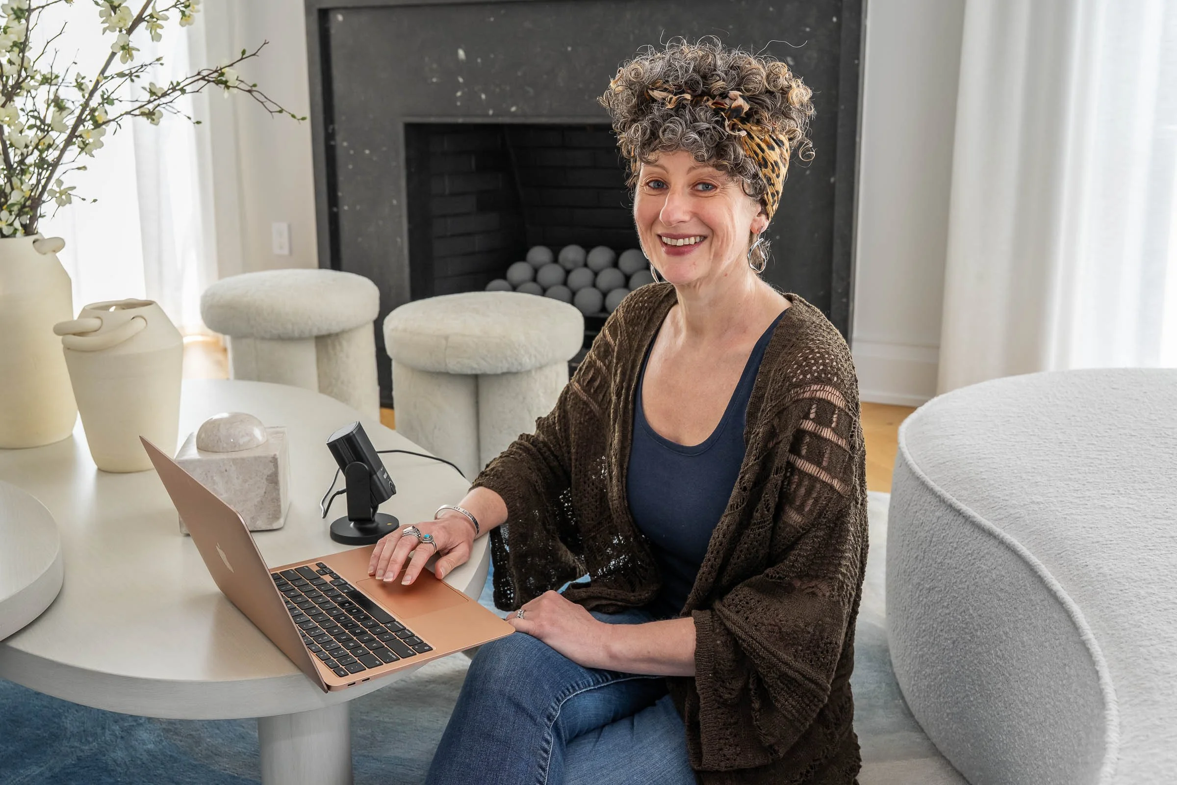 A woman with curly hair wearing a leopard print headscarf and a brown knitted cardigan sits at a white round table with a rose gold laptop in front of her. The background includes a black fireplace with white decorative balls, white curtains, and modern white furniture.
