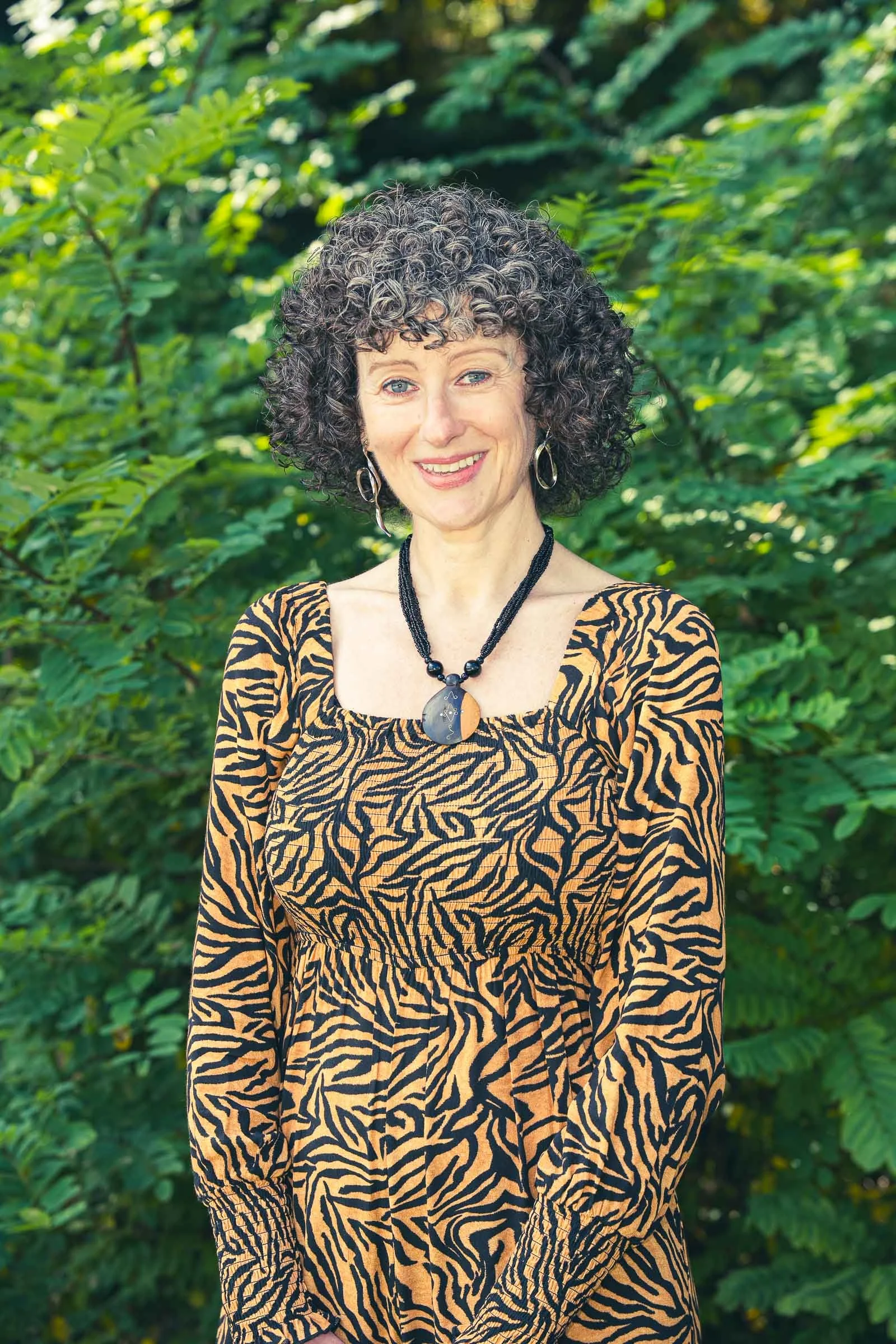 A woman with curly black and gray hair, wearing a tiger-print dress, earrings, and a necklace, standing outdoors with green foliage in the background and smiling at the camera.