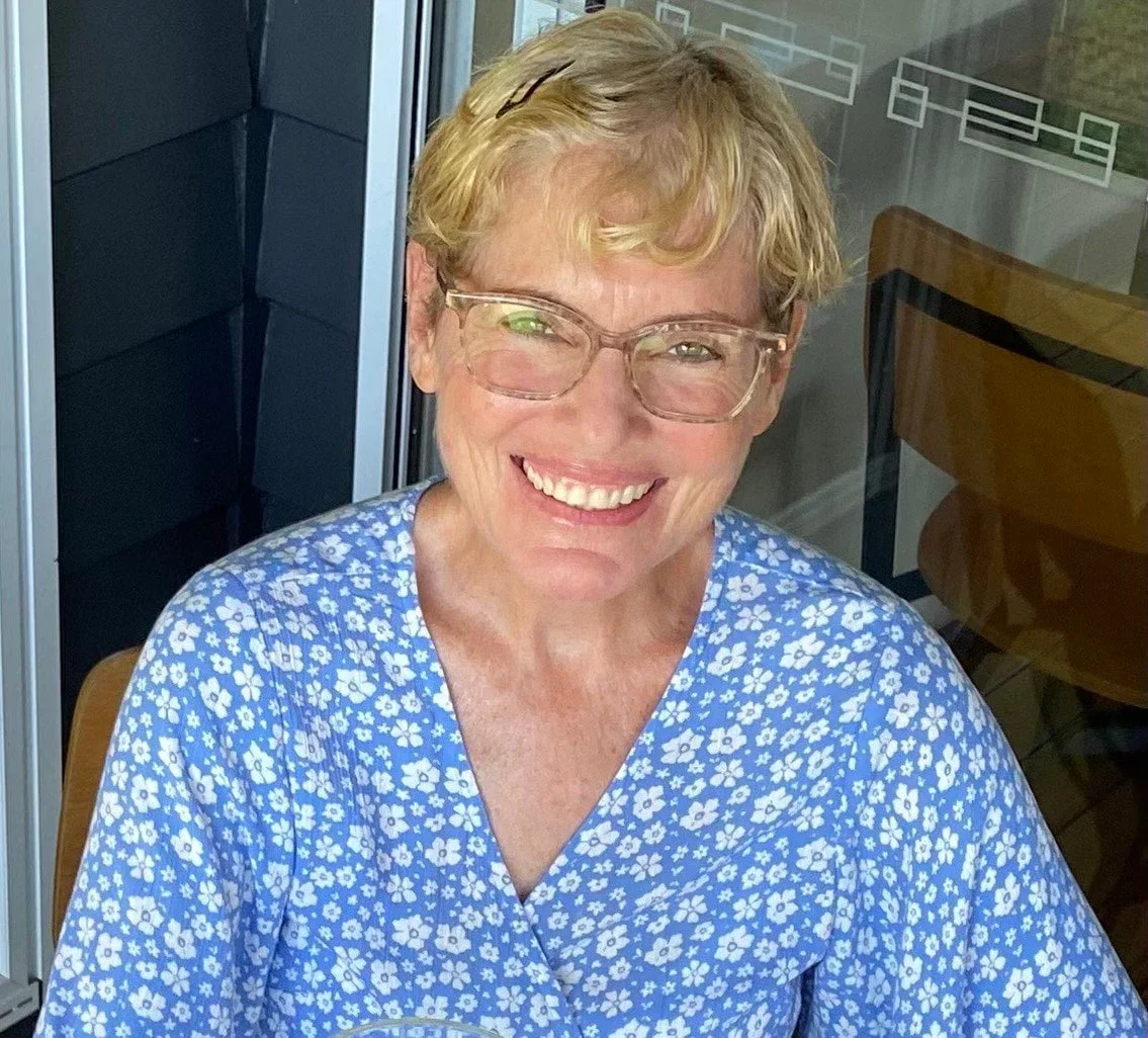A smiling woman with short blonde hair, wearing glasses and a blue floral dress, sitting outdoors near a glass door with a wooden chair beside her.
