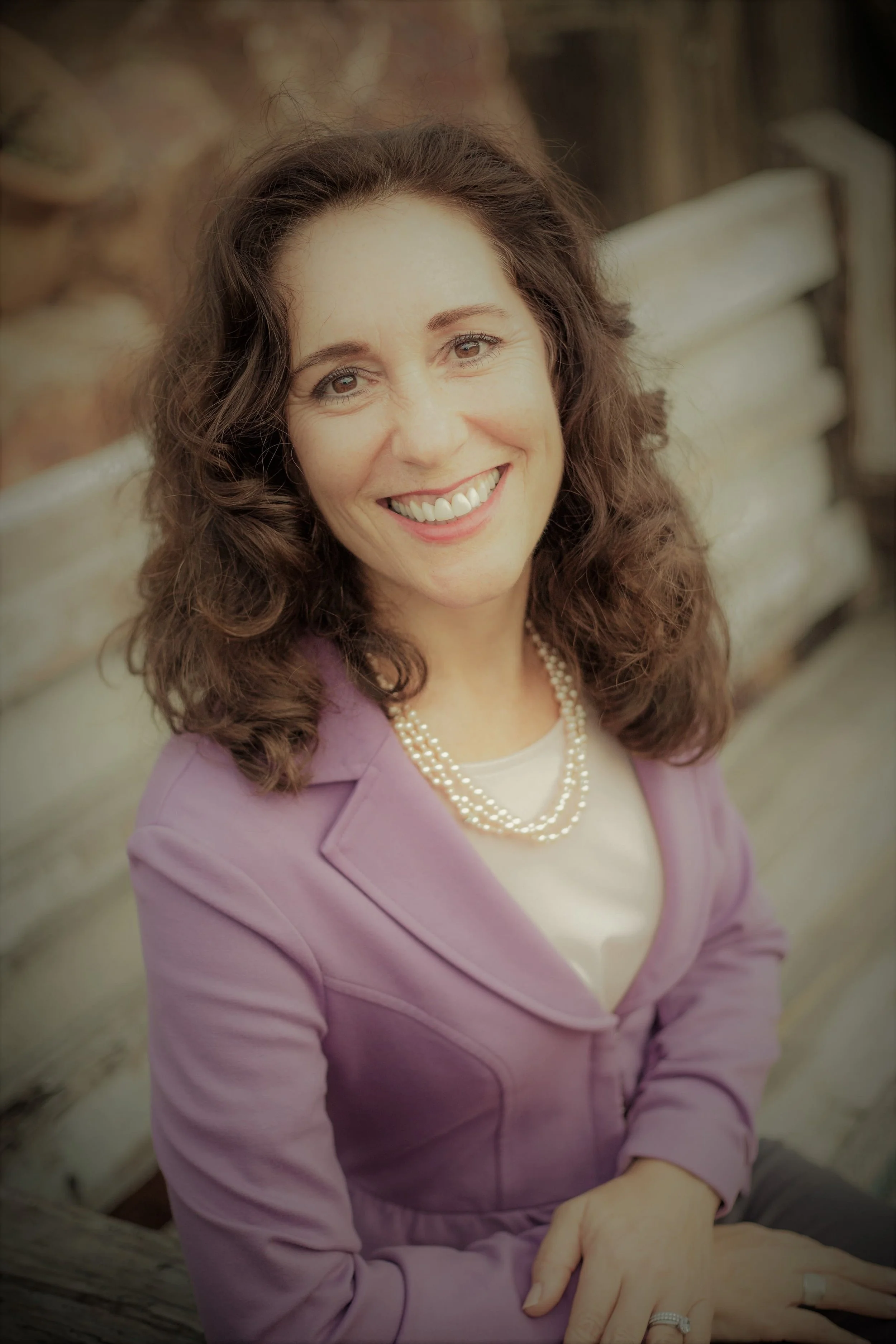 A woman with curly brown hair and a bright smile, wearing a purple blazer, a pearl necklace, and a white top, sitting on a weathered wooden bench outdoors.
