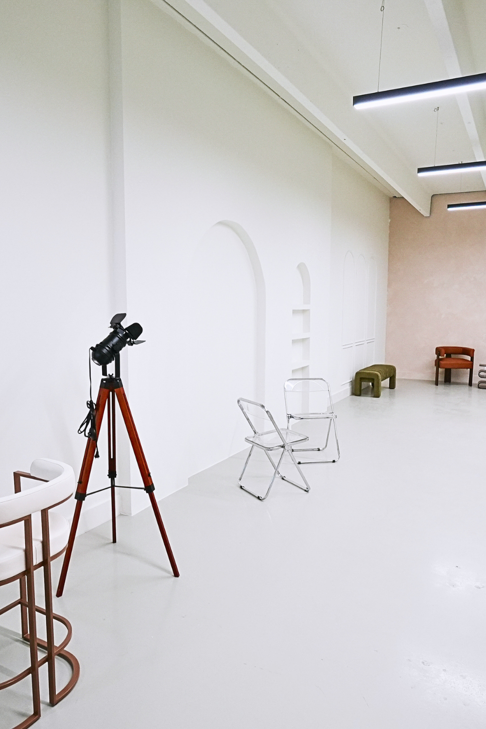 Empty art gallery with white walls, a white floor, and modern furniture, including a white chair, a green bench, and a brown chair, with a camera on a tripod.
