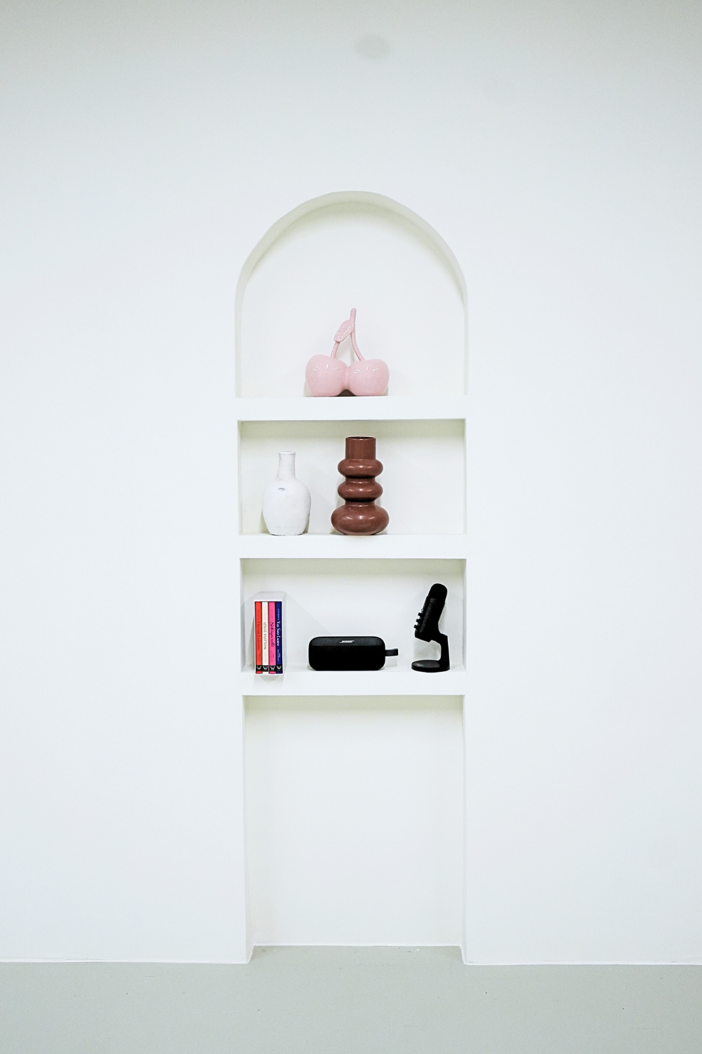 White wall with built-in niches displaying pink cherries sculpture, white and brown vases, black gadget, and a small black item.