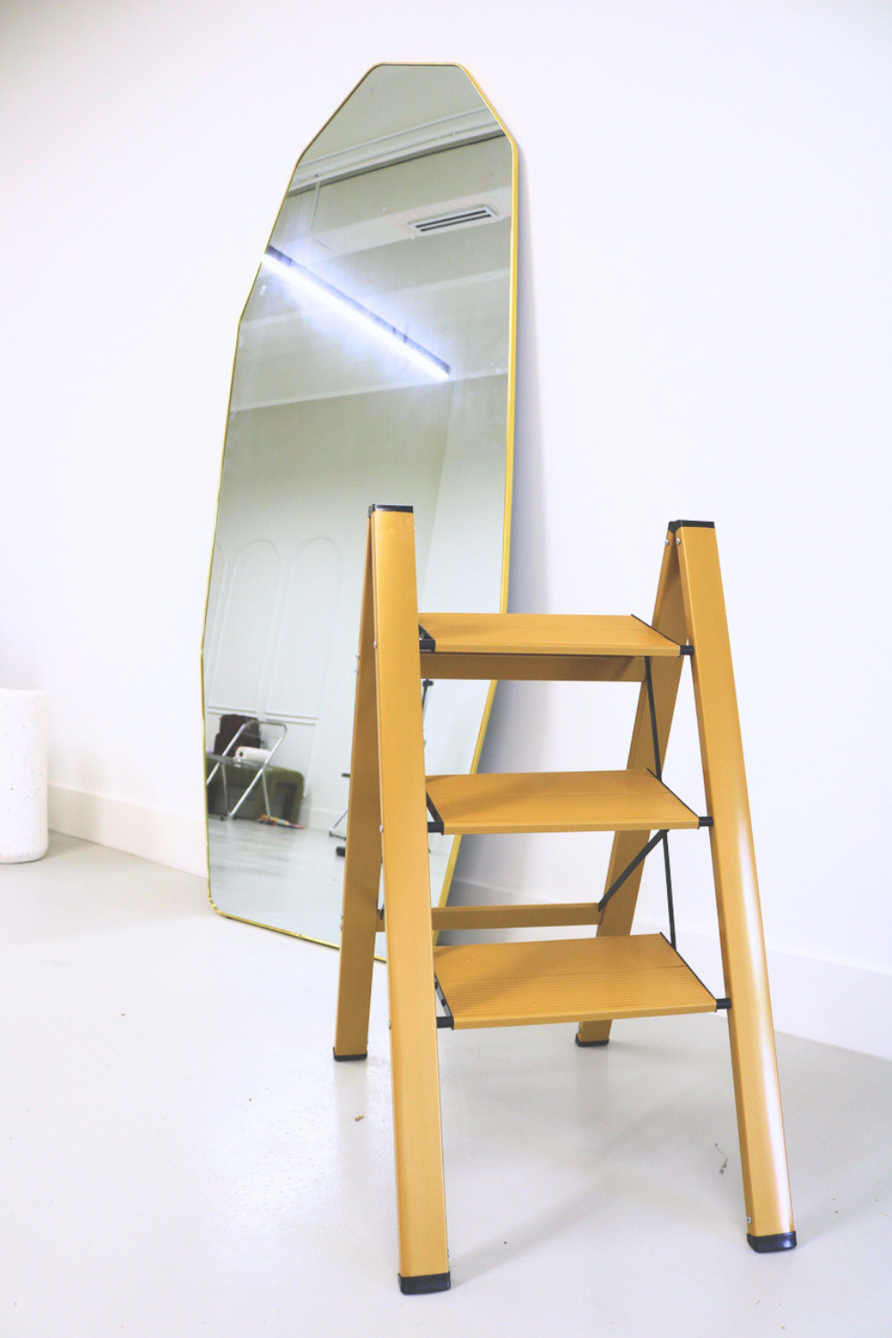 A gold-colored step ladder in front of a full-length mirror in a white room.
