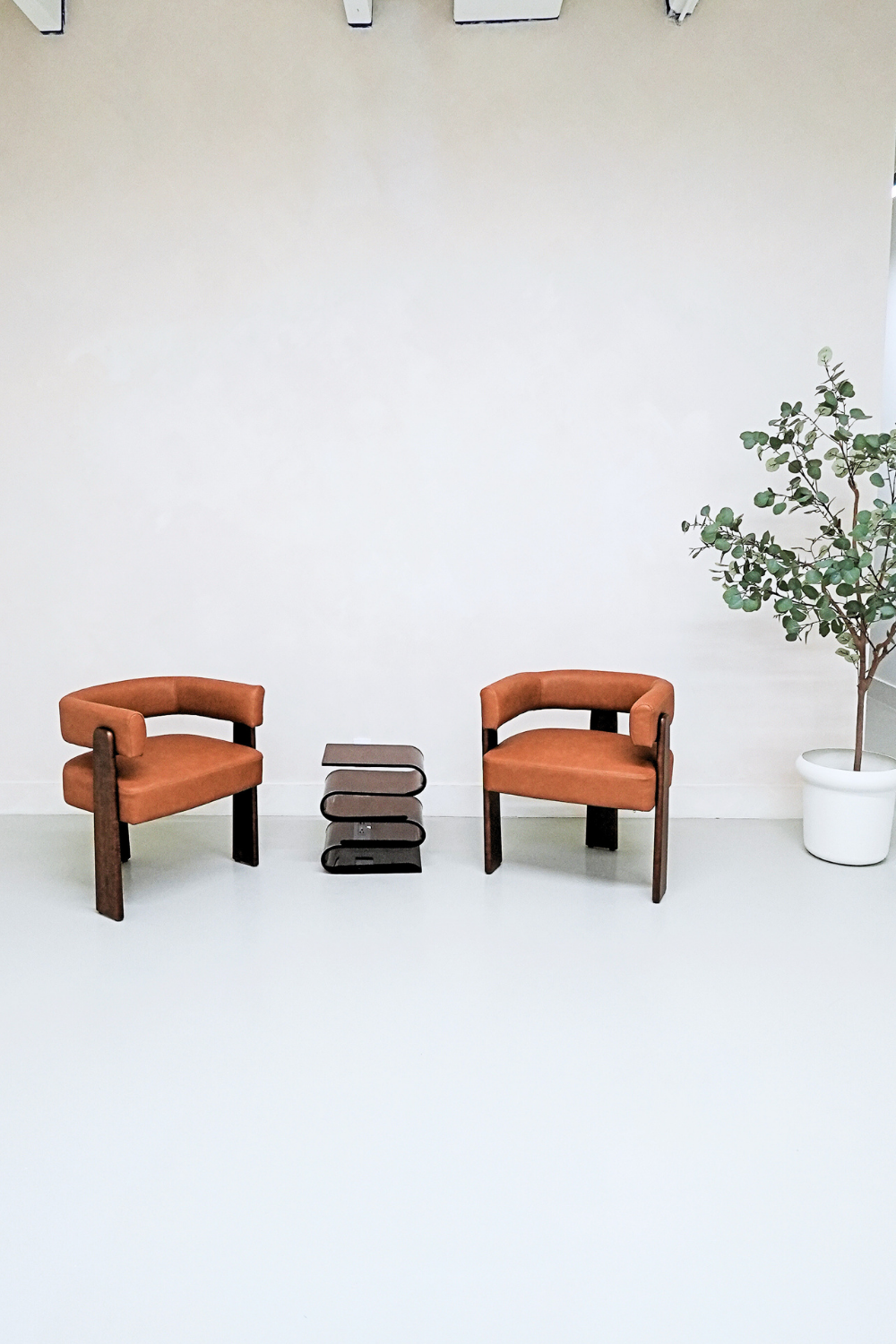 Two orange armchairs with wooden legs facing each other in a minimalist room with a white wall and light-colored floor, with a small glass table between them and a tall potted plant to the right.