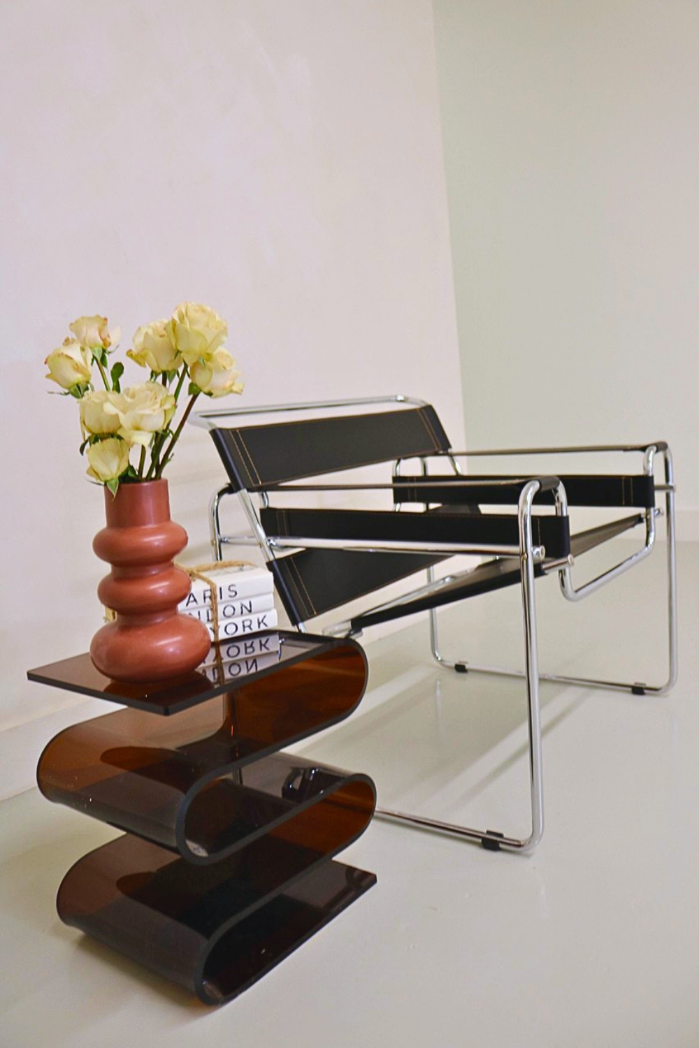 Interior scene with a black modern chair, a wooden side table, a pink vase with cream roses, and stacked books.