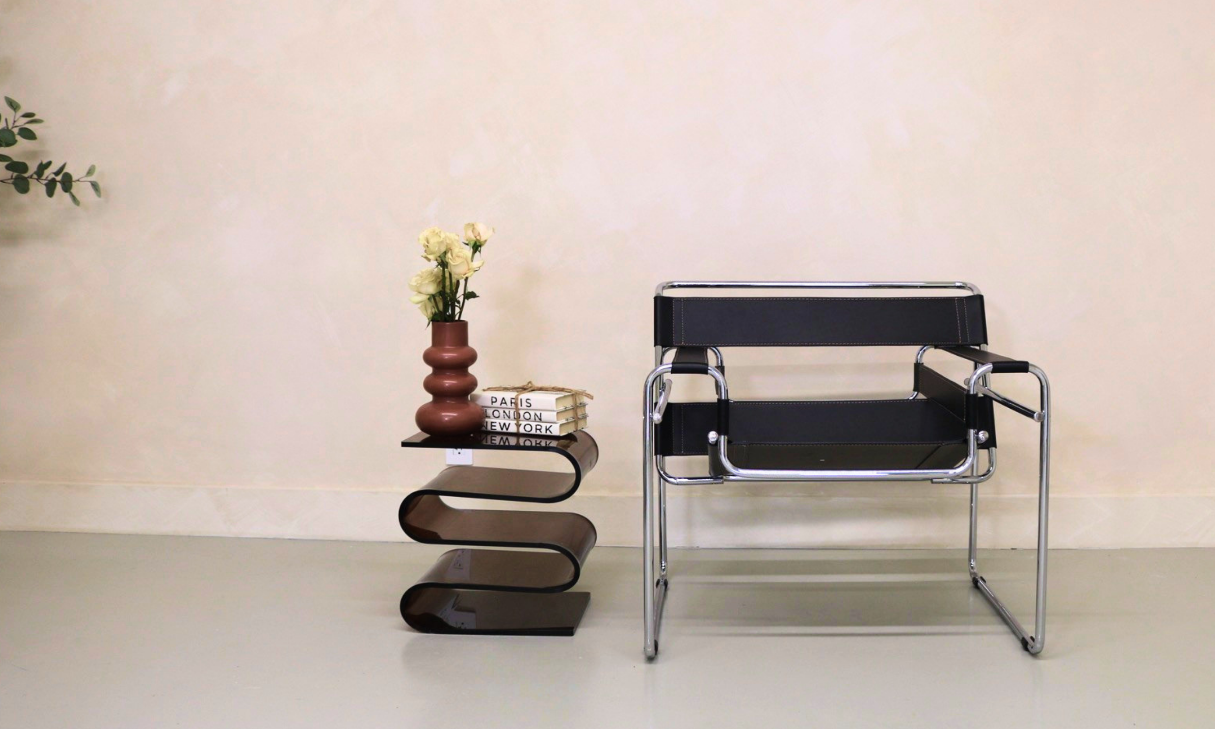 A contemporary interior with a black side table holding a brown vase with white flowers, a stack of books, and a modern black lounge chair with a chrome frame against a plain light wall.