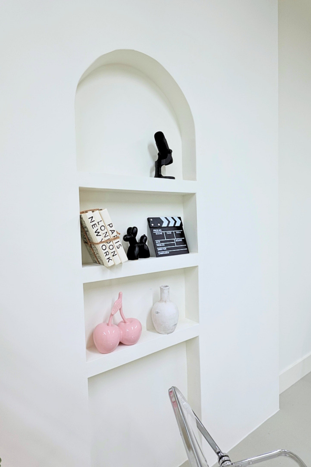 Decorative built-in wall shelves with black microphone, vintage books, black dog figurines, a papercraft clapperboard, a pink cherry-shaped ceramic decoration, and a white vase.