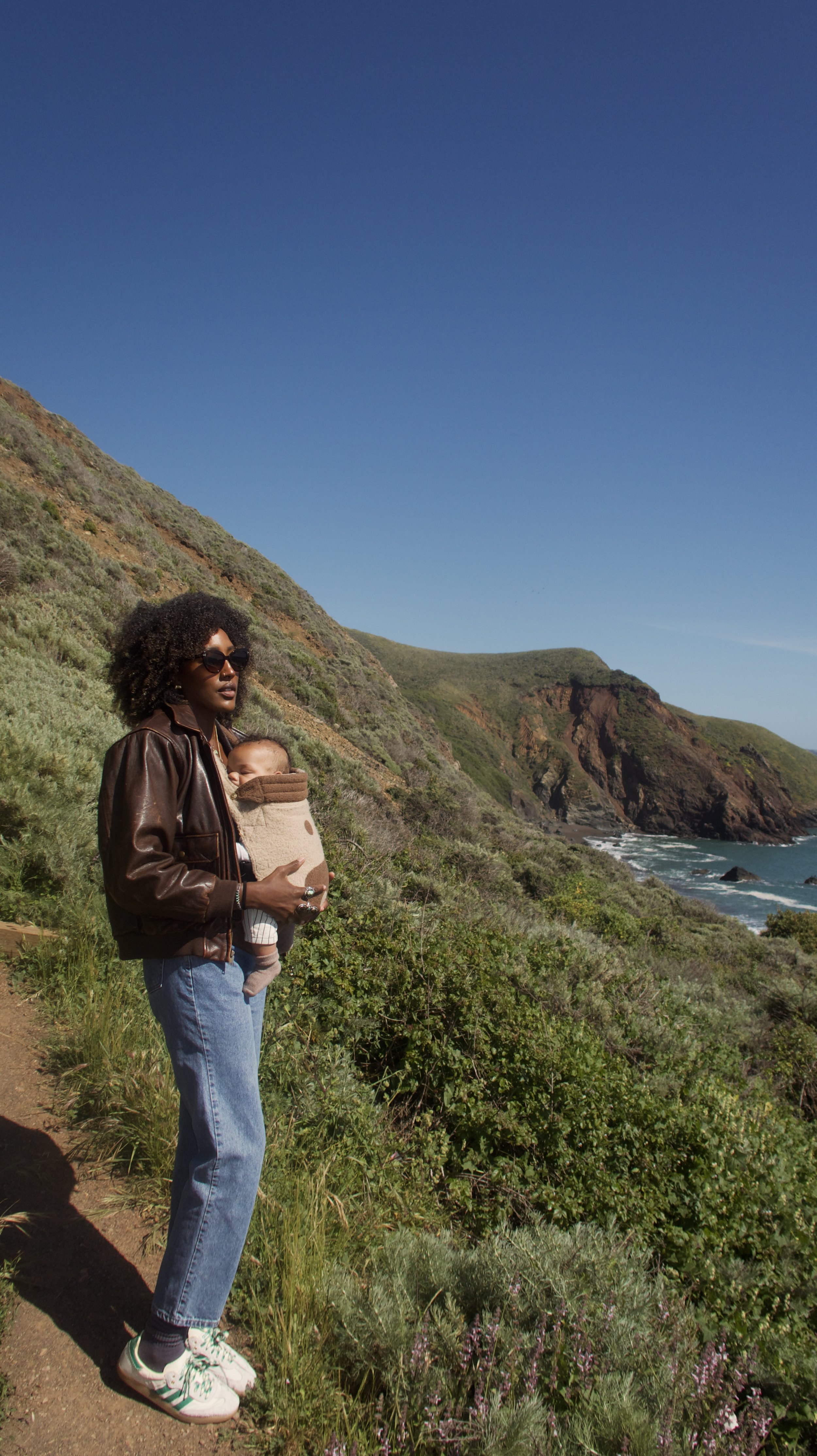 Woman with curly hair, wearing sunglasses, leather jacket, jeans, and sneakers, carrying a baby in a brown baby carrier, standing on a coastal trail with green hills and ocean in the background.