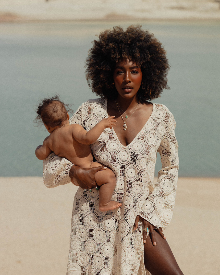 A woman with dark skin and curly hair, wearing a beige crochet dress, holding a baby with darker skin and curly hair, near a body of water on a sandy beach.