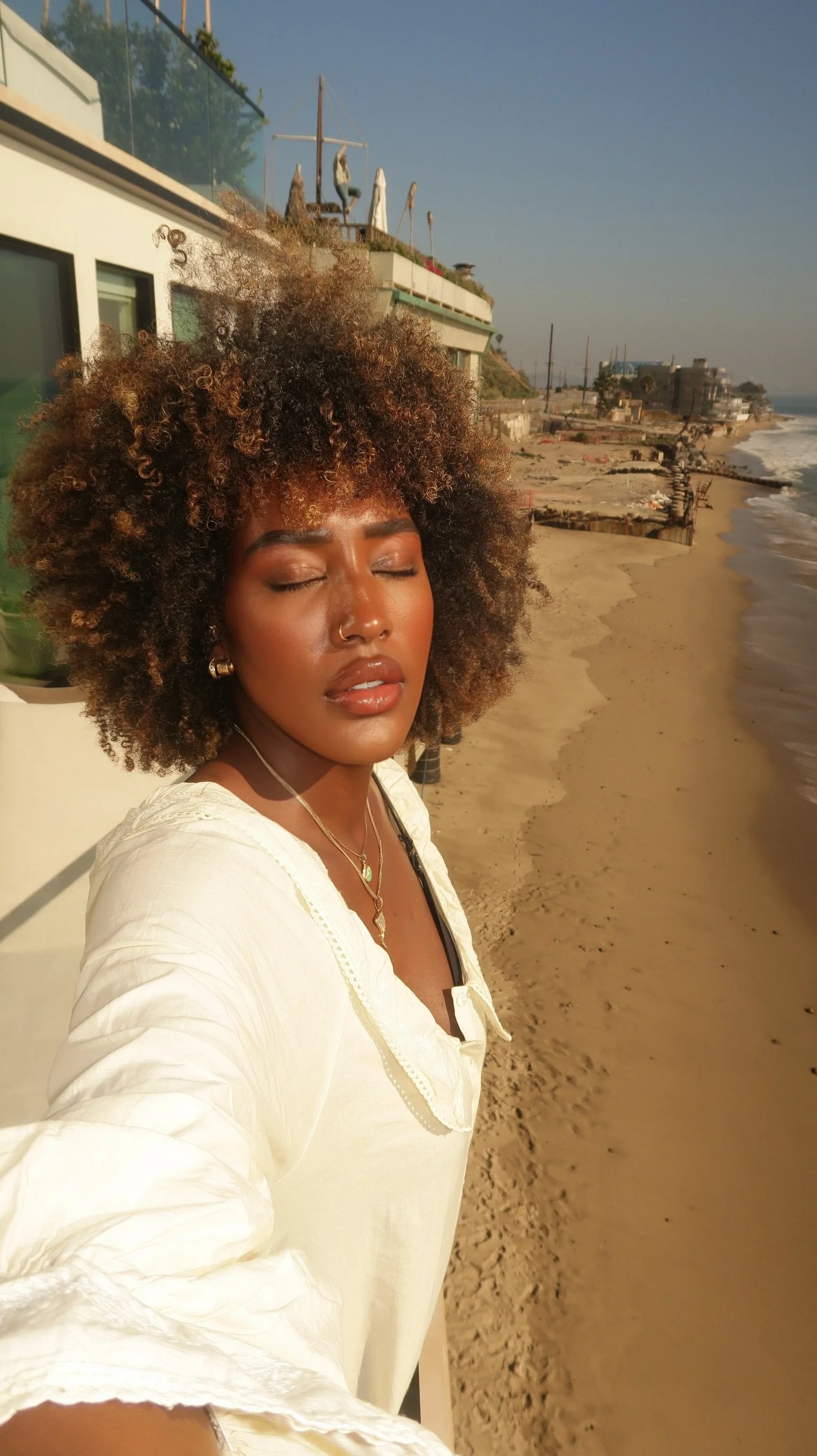 A woman with curly hair taking a selfie on a beach, with her eyes closed and sun shining on her face, sunset or sunrise lighting.