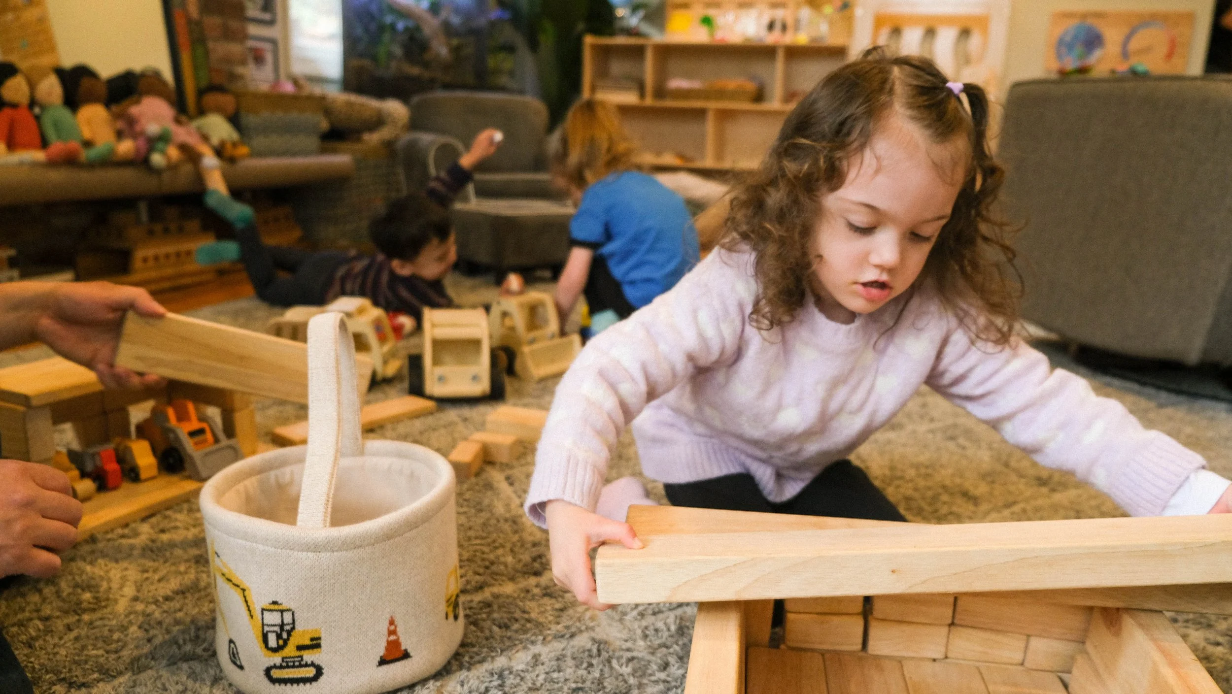 A young girl with curly hair playing with wooden blocks on a carpeted floor in a playroom. In the background, there are other children playing, a couch with plush toys, and shelves with toys and books.