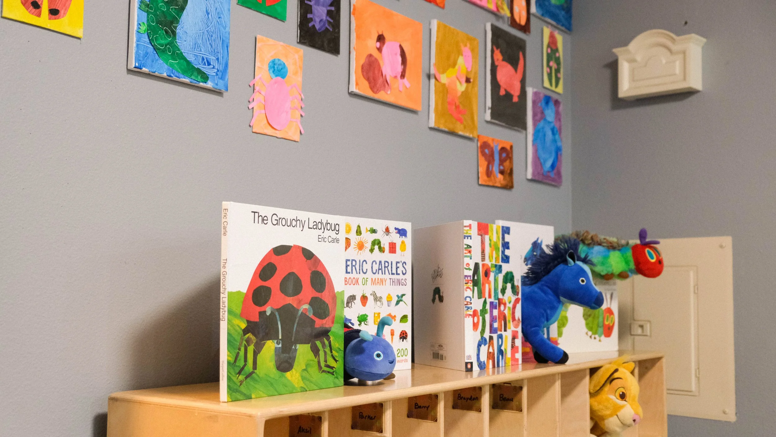 Children's books displayed on a wooden shelf with a wall of colorful animal artwork in the background. The books include titles about ladybugs and various animals, with plush animal toys on the shelf.