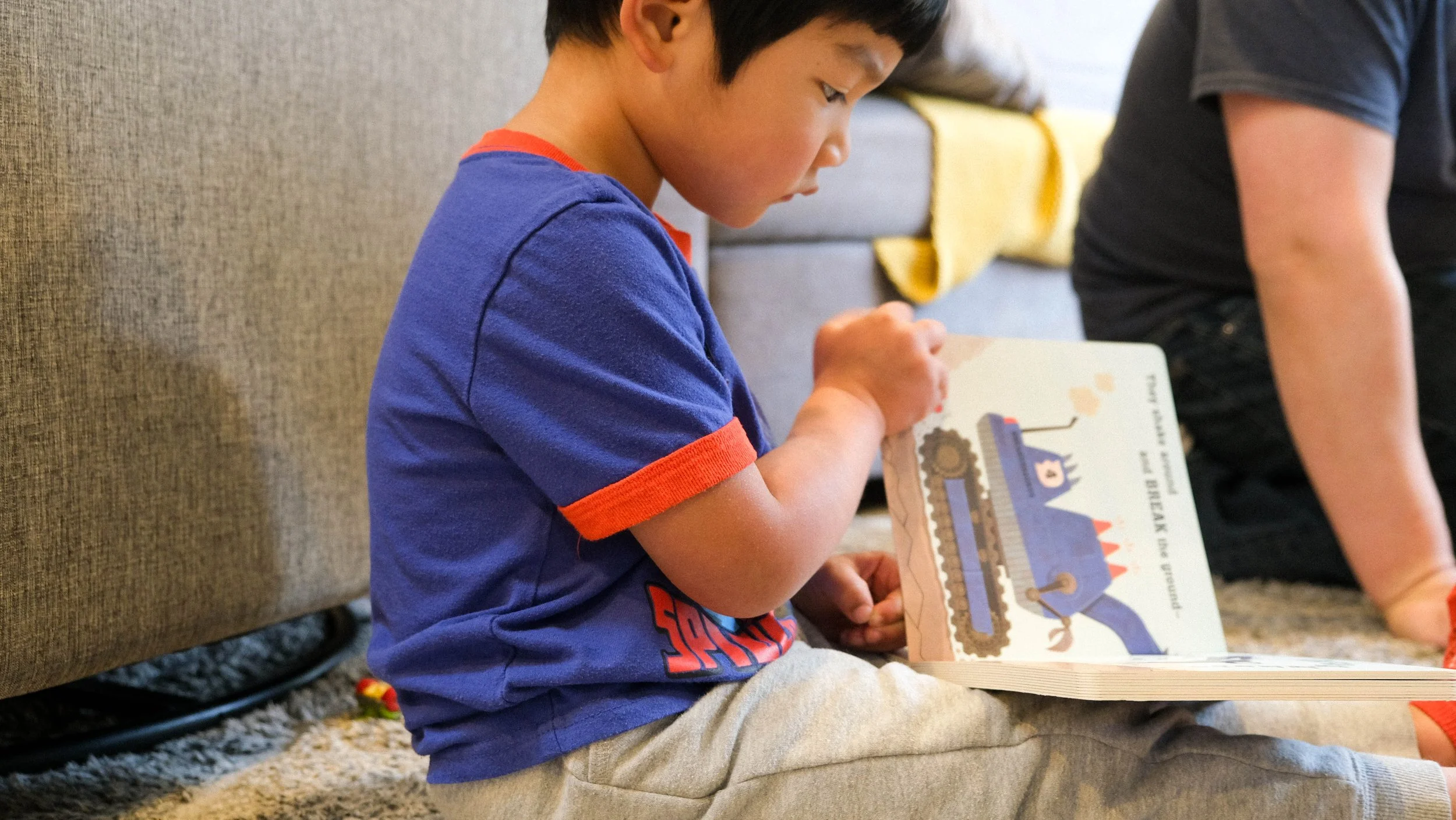 A young boy with black hair sitting on a carpeted floor, reading a colorful picture book about a train, with a beige couch and a yellow blanket in the background.