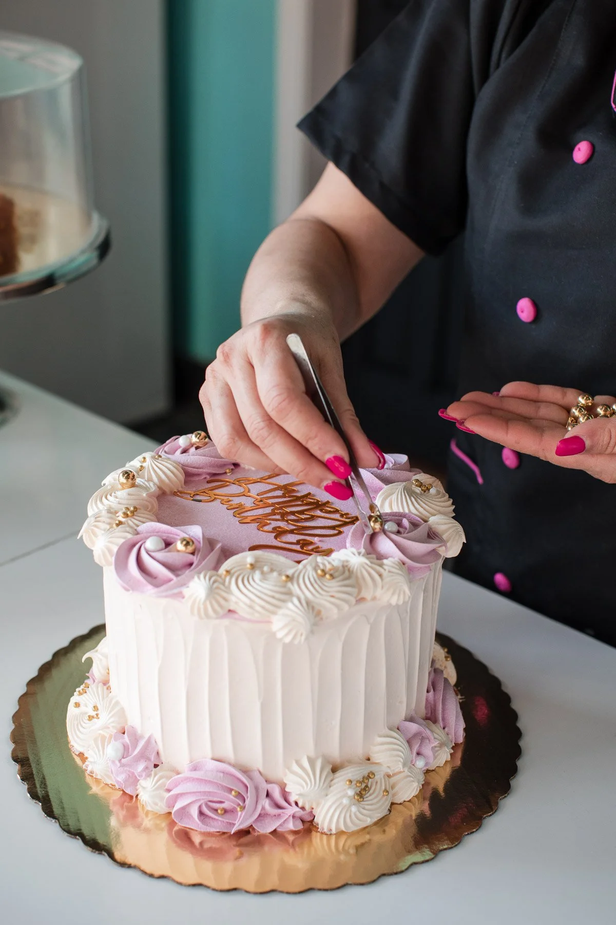 A person is decorating a pink and white birthday cake with piped frosting and gold accents. The individual is using tweezers to place a gold decorative element on the cake. The cake is on a reflective, gold-colored base, and the person has pink nail polish and is wearing a black shirt with pink buttons.
