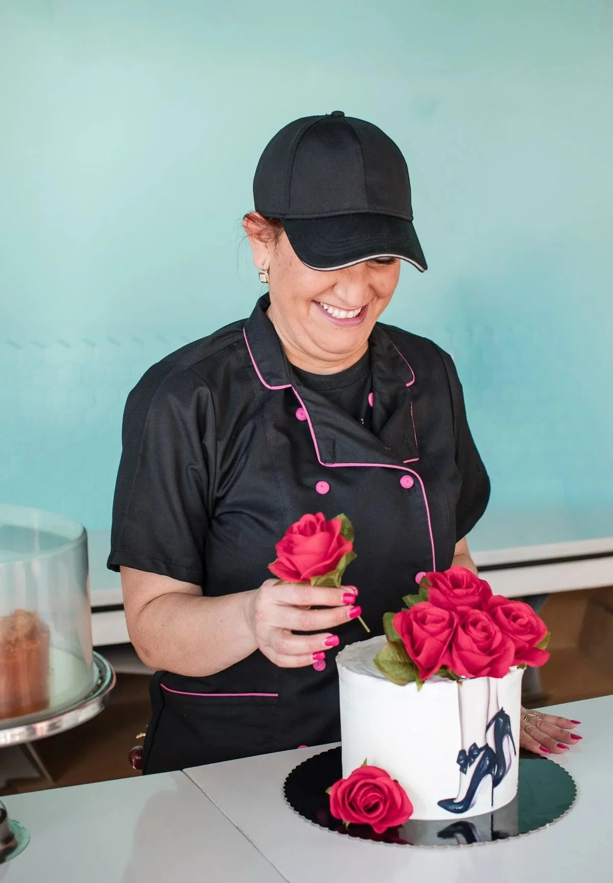 A woman in a black chef uniform with pink piping and a black cap is smiling and decorating a cake with pink roses.
