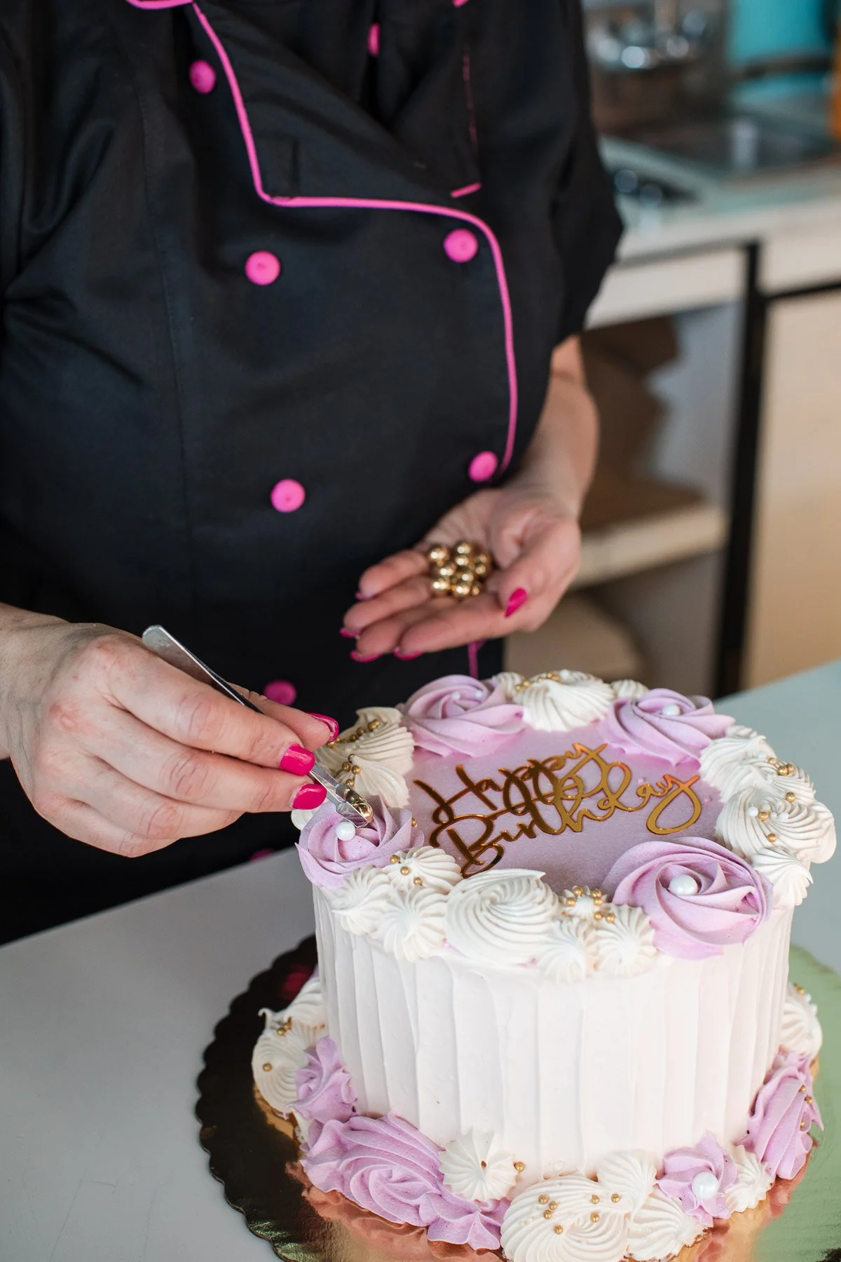 Person in a black chef's coat with pink trim decorating a birthday cake with a piping tool. The cake is white with pink and white icing roses and gold pearl decorations, topped with a "Happy Birthday" gold script topper.