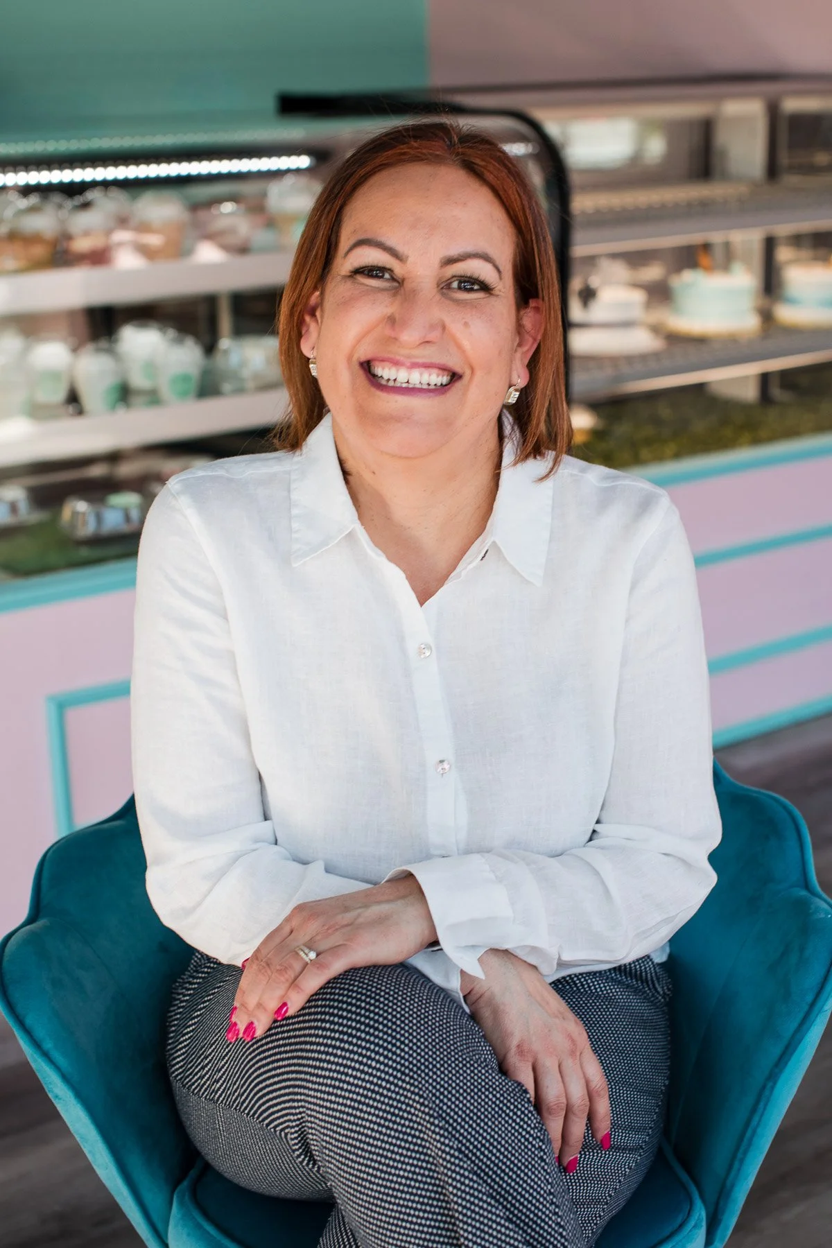 A woman with red hair smiling, sitting on a blue chair in front of a display with cakes and desserts.