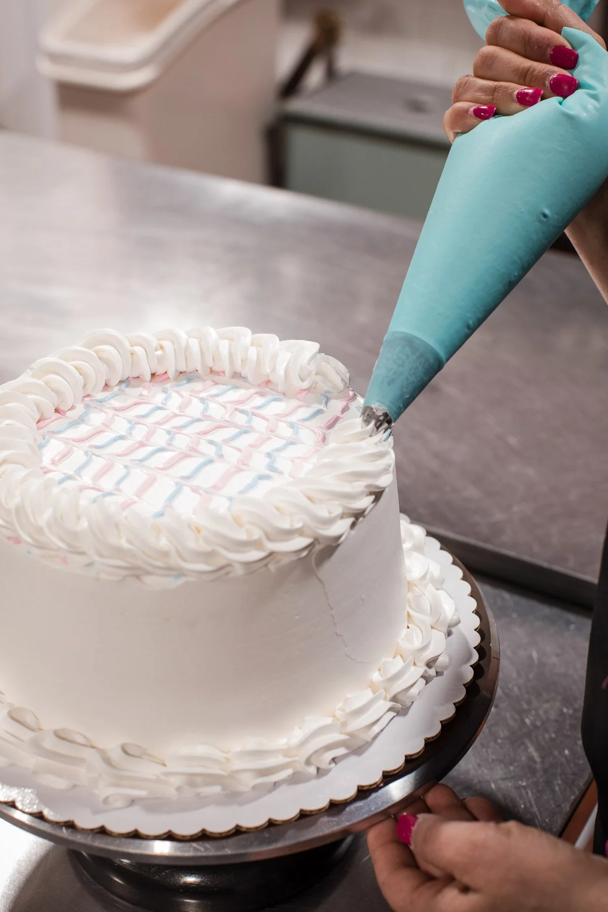 A person with pink painted nails is using a piping bag to decorate a large white cake with white frosting, creating a decorative border on top and around the sides of the cake.